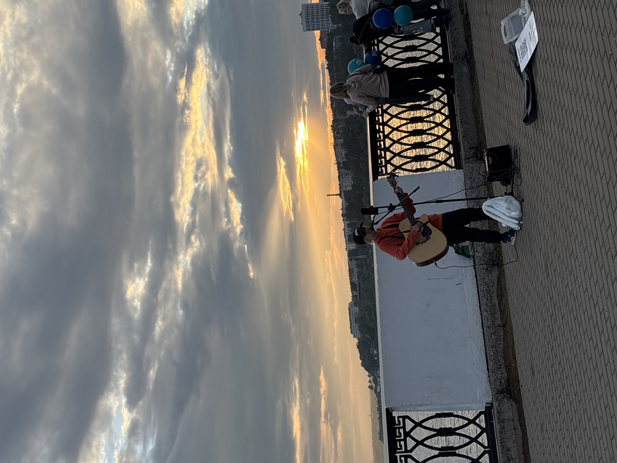The image captures a sunset over the Izh River in Izhevsk, Russia. A street musician, wearing an orange hoodie and a black baseball cap, plays an acoustic guitar on the embankment. In front of him are a microphone stand, a small amplifier, and a white cloth bag. To his right, on the other side of an ornate black metal fence, a small group of people are gathered, some of whom are holding balloons. The sky is dramatic, with rays of sunlight breaking through a cloudy expanse. The embankment itself is paved with brickwork. In the distance, across the river, is a cityscape with buildings and trees.
