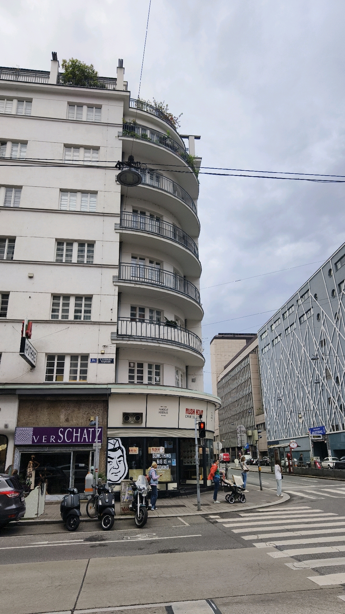 The image depicts a street corner in Vienna, Austria, with a prominent white modernist apartment building featuring curved balconies. The building's facade is punctuated by rows of windows. At street level, a storefront displays the name "VER SCHATZ" with mannequins visible inside. Adjacent to it is another shop with signage that includes "RUSH HOUR" and "CHINESE FOOD." A red traffic light is illuminated above the intersection. Several parked motorcycles and scooters are present on the sidewalk. A pedestrian crossing spans the street. Further down the street, modern buildings with geometric facades are visible. The sky is overcast, indicating a cloudy day. A few people are visible on the sidewalk, including a woman pushing a stroller and another individual standing near a traffic light.