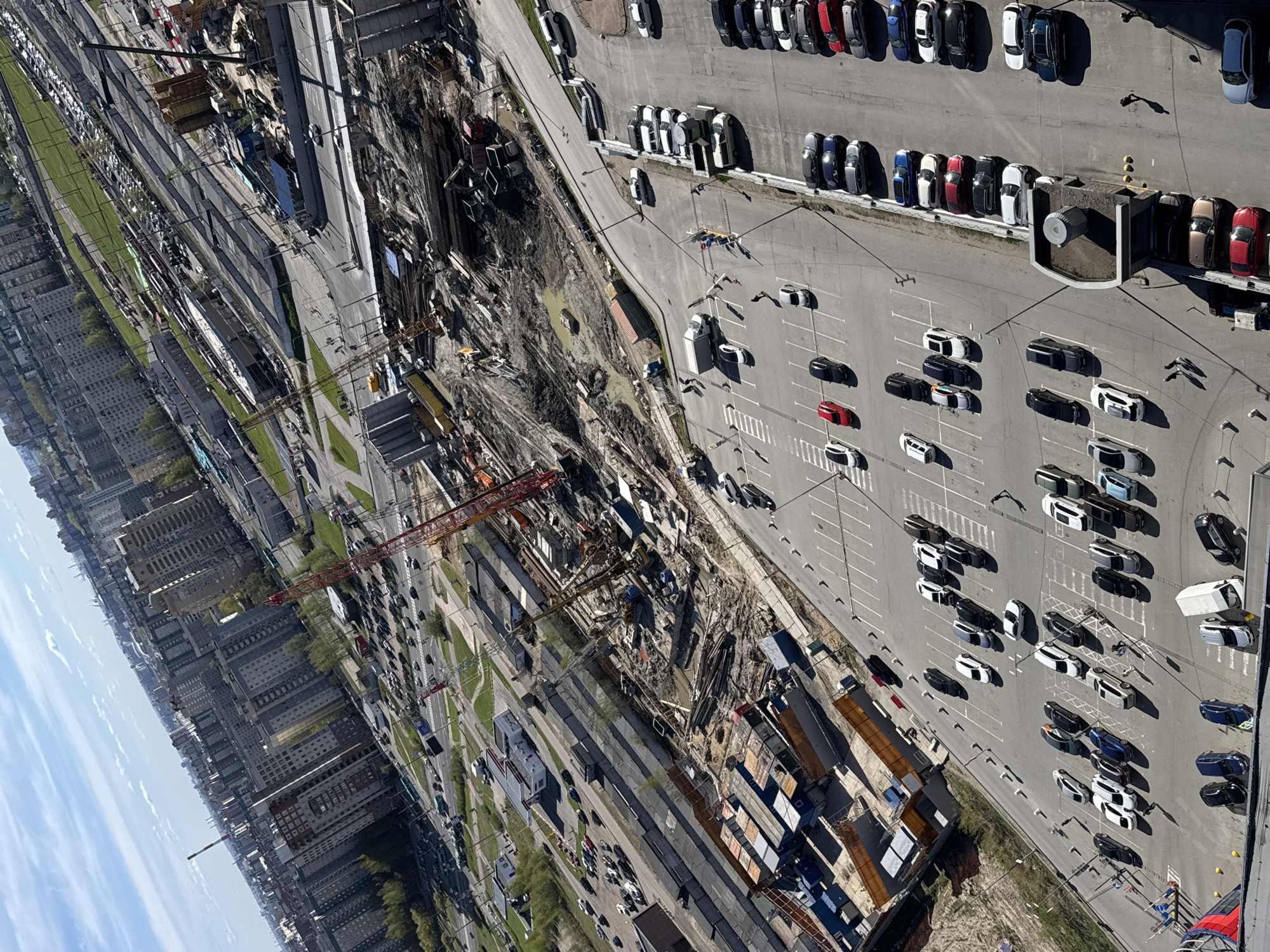 An aerial view shows ongoing construction adjacent to a parking lot in Saint Petersburg, Russia. The parking lot is densely packed with vehicles, and a large pit reveals the construction site with various building materials and construction equipment present. Buildings are visible in the background.