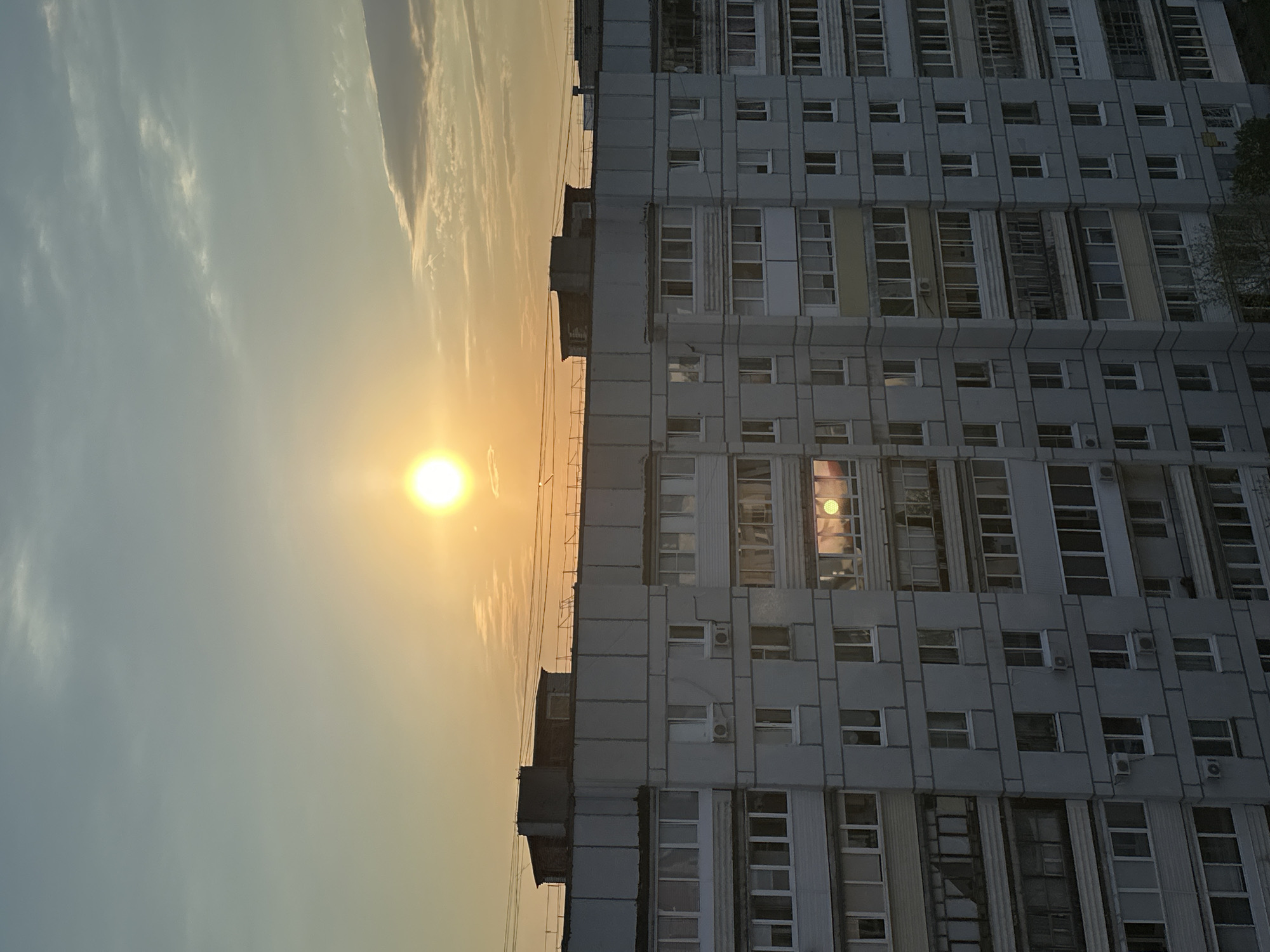 The image shows an apartment building located in Chaikovsky, Russia, at sunset. The building is multi-story, constructed of concrete, and features repetitive window arrangements. The setting sun illuminates the sky with a warm glow, reflecting off some of the building's windows. The sky has a hazy, slightly overcast appearance. There are no people visible in the frame.