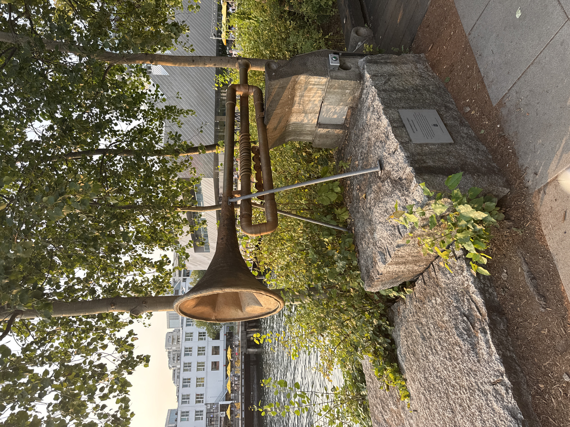 The image depicts an outdoor art installation situated by a body of water, likely the Burrard Inlet, in North Vancouver, Canada. The artwork, which appears to be made of rusted metal pipes and a large horn-like structure, is mounted on a stone base. It is surrounded by greenery and positioned near trees. In the background, modern buildings and a boardwalk with yellow umbrellas are visible, suggesting a public waterfront area. The lighting indicates it is late afternoon or early evening. No people are prominently featured in the immediate vicinity of the artwork. The overall scene is one of an urban waterfront park with a prominent sculptural element.