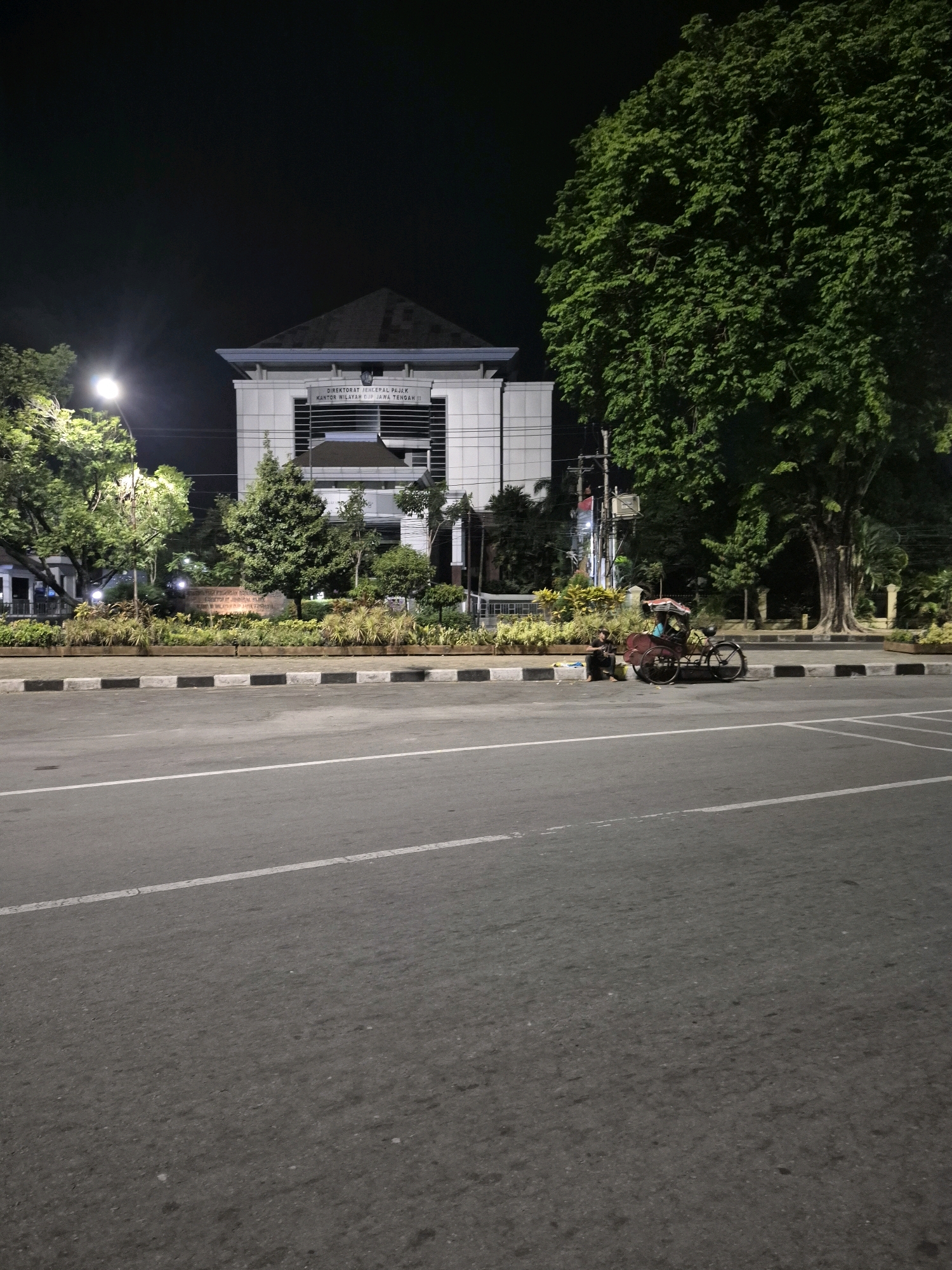 The image depicts an outdoor scene at night, likely in the city of Surakarta, Indonesia, given the text on the building which appears to be in Indonesian. In the foreground, a paved road with white lane markings stretches across the frame. To the left of the road, a sidewalk is visible, bordered by a curb with black and white patterns. Beyond the sidewalk, there is a landscaped area with various plants and trees.
Dominating the background is a modern, multi-story building with a light-colored facade and a dark, angled roof. Text is visible on the building, suggesting it is a government or administrative office. To the right of the building, a large, mature tree with dense green foliage stands prominently.
On the sidewalk, near the landscaped area and in front of a large tree, a rickshaw is parked. A person is seated beside the rickshaw, facing away from the camera. Another person is seated on the ground, near the sidewalk, looking towards the rickshaw. The lighting suggests that the area is illuminated by streetlights and lights from the building, casting shadows and highlighting certain elements of the scene. The overall atmosphere is quiet and still, consistent with a late-night setting.