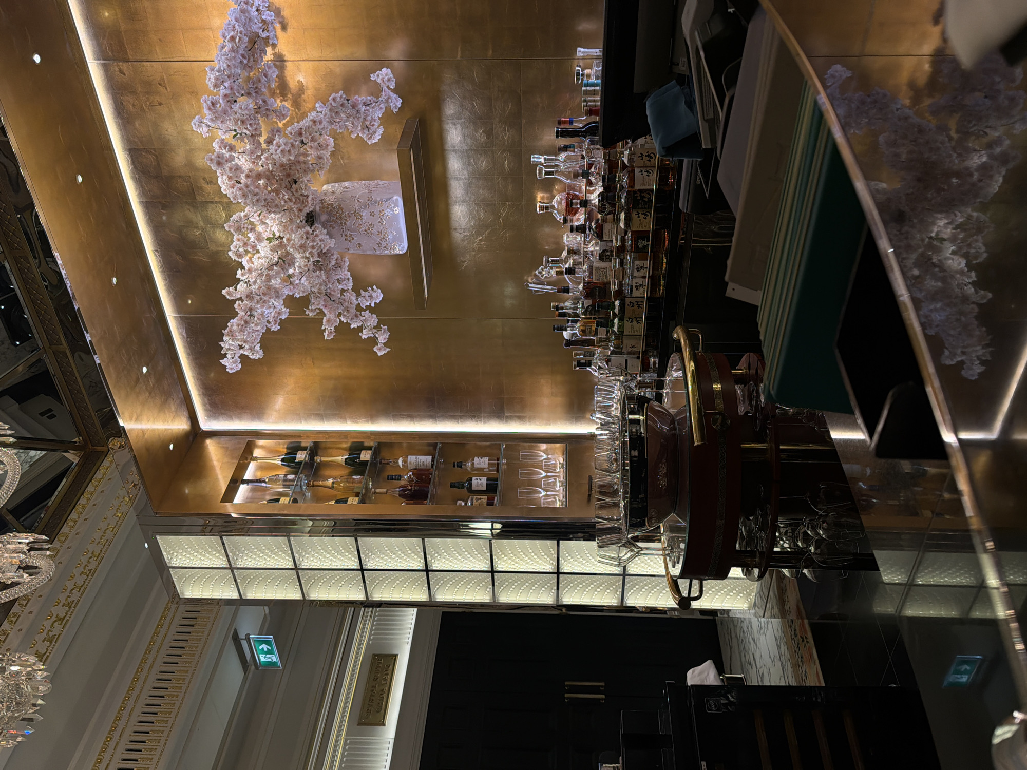 The image captures the interior of a luxurious bar, possibly located in London, United Kingdom. The scene showcases a blend of opulence and contemporary design.
The upper portion of the frame features a gold-textured wall adorned with a decorative floral arrangement resembling cherry blossoms. Below this, a well-stocked bar displays an array of liquor bottles and glassware, indicating a focus on mixology.
The lower section reveals a hallway or foyer with ornate architectural details, including a chandelier and decorative molding, consistent with a high-end establishment. An illuminated "Exit" sign indicates an emergency escape route. The floor is highly polished, reflecting the surrounding light and adding to the overall sense of refinement.
