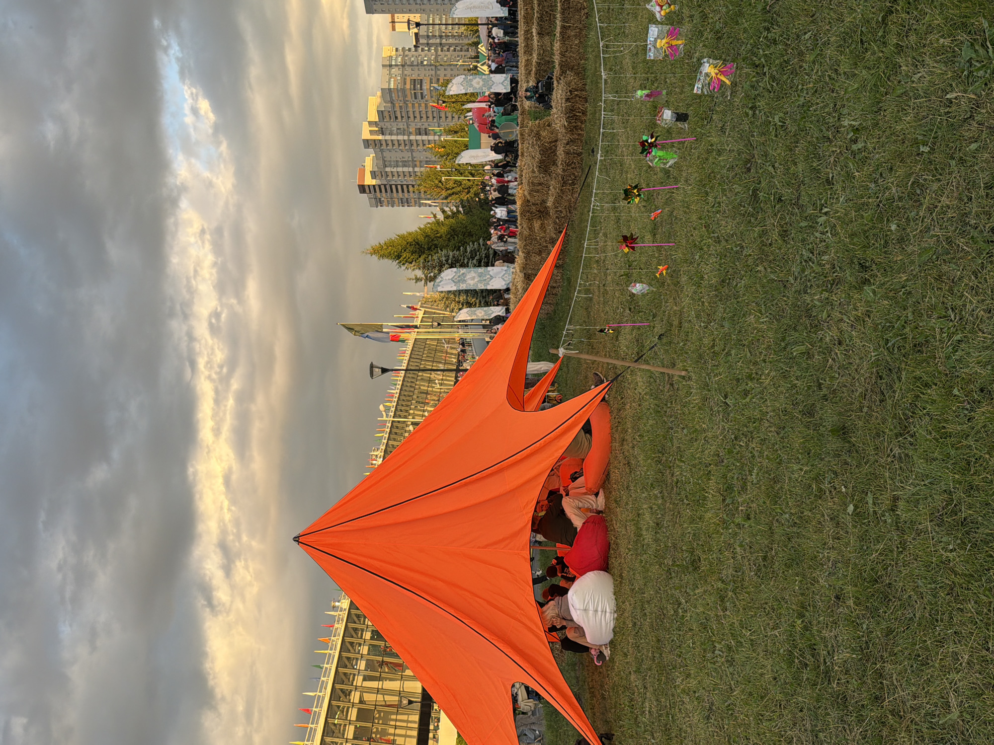 In an outdoor urban park setting, a group of people are gathered under a large, bright orange, star-shaped canopy tent. The tent is set up on a grassy area, with several large, white beanbag chairs and smaller red and orange beanbag chairs scattered within its shaded interior. Some individuals are seated on these chairs, engaged in conversation or relaxing. In the background, a crowd of people are milling about, with flags and banners visible. A structure that appears to be a modern building with multiple levels and windows is to the left, adorned with numerous flags. Further in the distance, modern, multi-story residential buildings are visible against a cloudy sky with patches of yellow and white light. In the foreground, a string of colorful, decorative pinwheels and small toys are hanging, swaying gently in the breeze. The scene suggests a public gathering or festival in a city park.