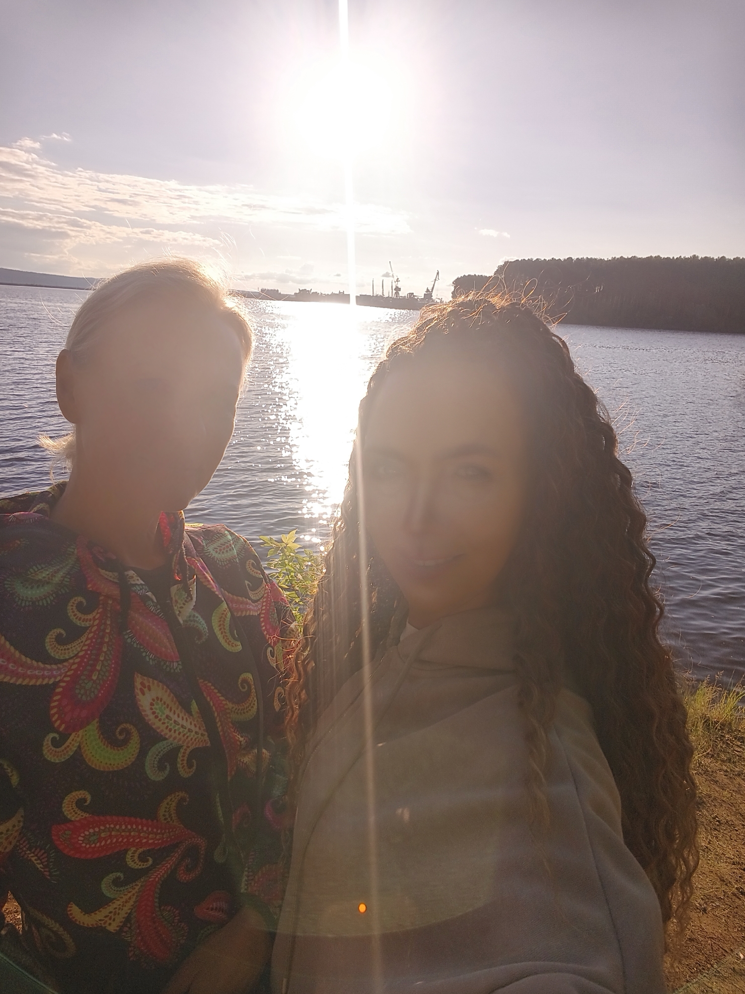 Two women are featured in a sun-drenched outdoor selfie, positioned against a scenic backdrop of a body of water during either sunrise or sunset. The bright sun is directly behind them, creating a strong backlight, prominent lens flare, and casting their faces largely into shadow.
The woman on the left has light-colored hair and is wearing a dark-colored top with a vibrant, intricate paisley pattern featuring shades of red, orange, green, and blue. Her facial features are mostly obscured by the shadow.
The woman on the right has long, dark, curly hair that cascades over her shoulder. She is wearing a light-colored, possibly beige or gray, top, which appears to be a hoodie. A subtle smile is discernible on her face despite the challenging lighting conditions.
Behind the women, a wide expanse of water reflects the intense sunlight, creating glittering patterns on its surface. Across the water, a distant shoreline reveals industrial structures, including several cranes, alongside a densely forested area. The immediate foreground where the women are standing shows a dry, grassy bank. There is no visible text in the image.