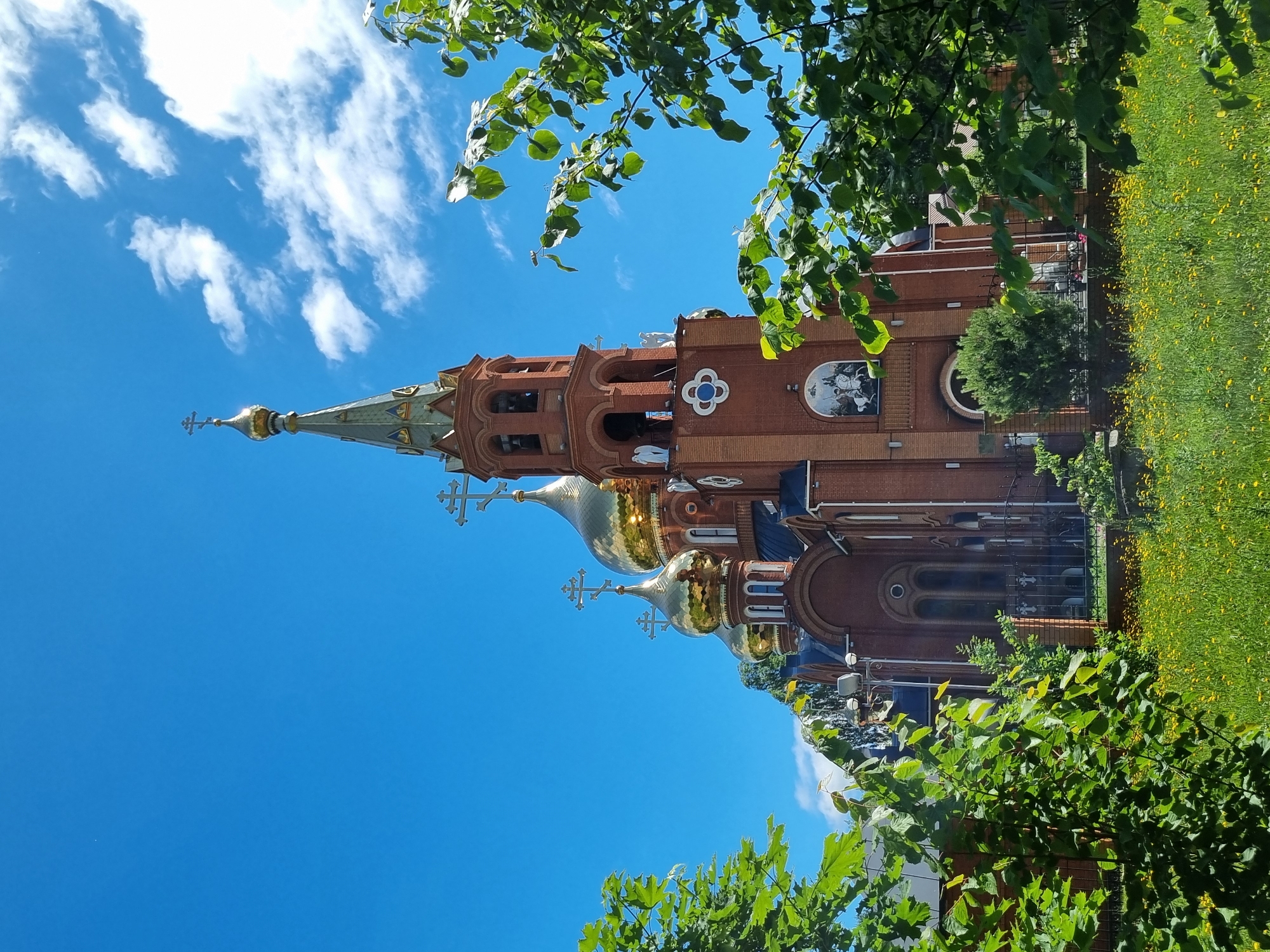 An Orthodox church with multiple golden domes and a tall bell tower made of red brick stands against a bright blue sky with scattered white clouds. The church has arched windows and decorative elements, including crosses atop the domes and a mural above an entrance. Lush green trees frame the church on the left and right, and a field of yellow wildflowers is visible in the foreground. The scene appears to be set in a park or garden. The church is the Church of the Kazan Icon of the Mother of God in Chaikovsky, Russia. There are no people visible in the image.