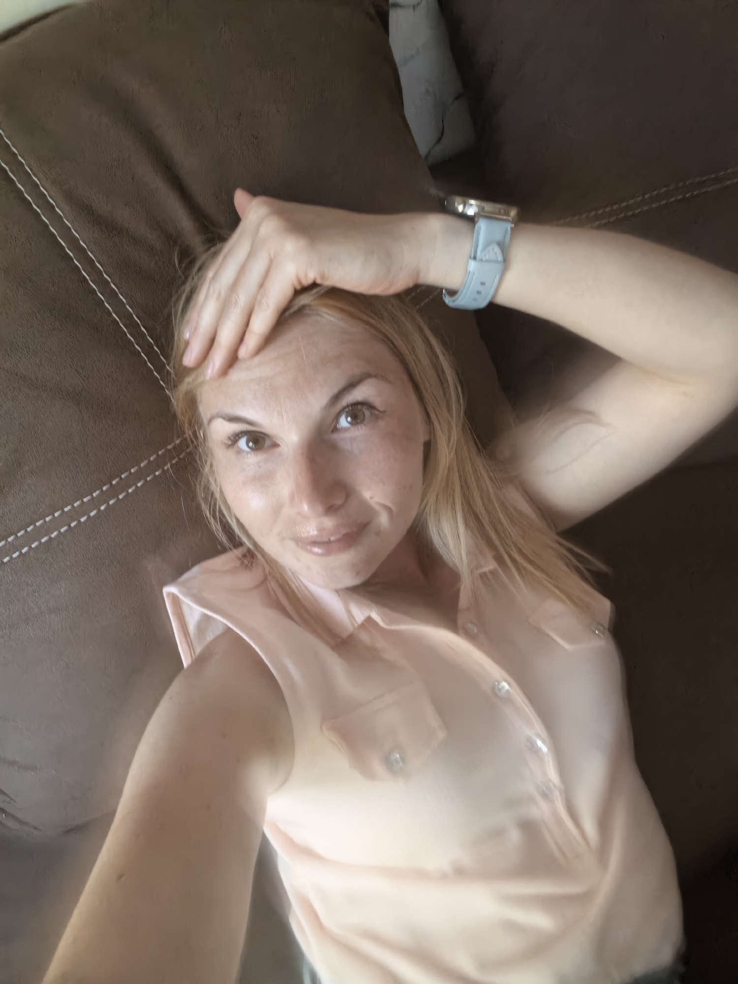 A woman lies on a brown sofa. She is wearing a light pink blouse with buttons and has a light blue watch on her wrist. She has blonde hair and is touching her forehead with her hand. The location is unknown.