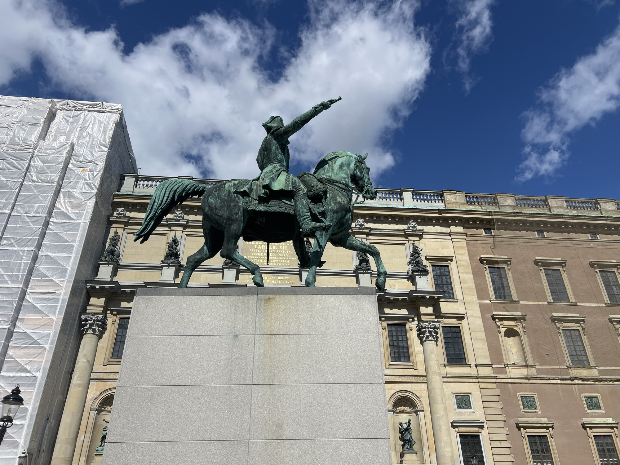 This image depicts the equestrian statue of King Charles XII (Karl XII) located in front of the Royal Palace in Stockholm, Sweden. The bronze statue, showing the king on horseback pointing forward, is mounted on a large granite pedestal. To the left of the statue, a portion of a building is covered in white scaffolding and sheeting, indicating ongoing construction or renovation. Behind the statue, the facade of the Royal Palace is visible, featuring classical architecture with numerous windows and decorative elements. The sky above is a deep blue with scattered white clouds. The perspective is from a low angle, looking up at the statue and the palace.