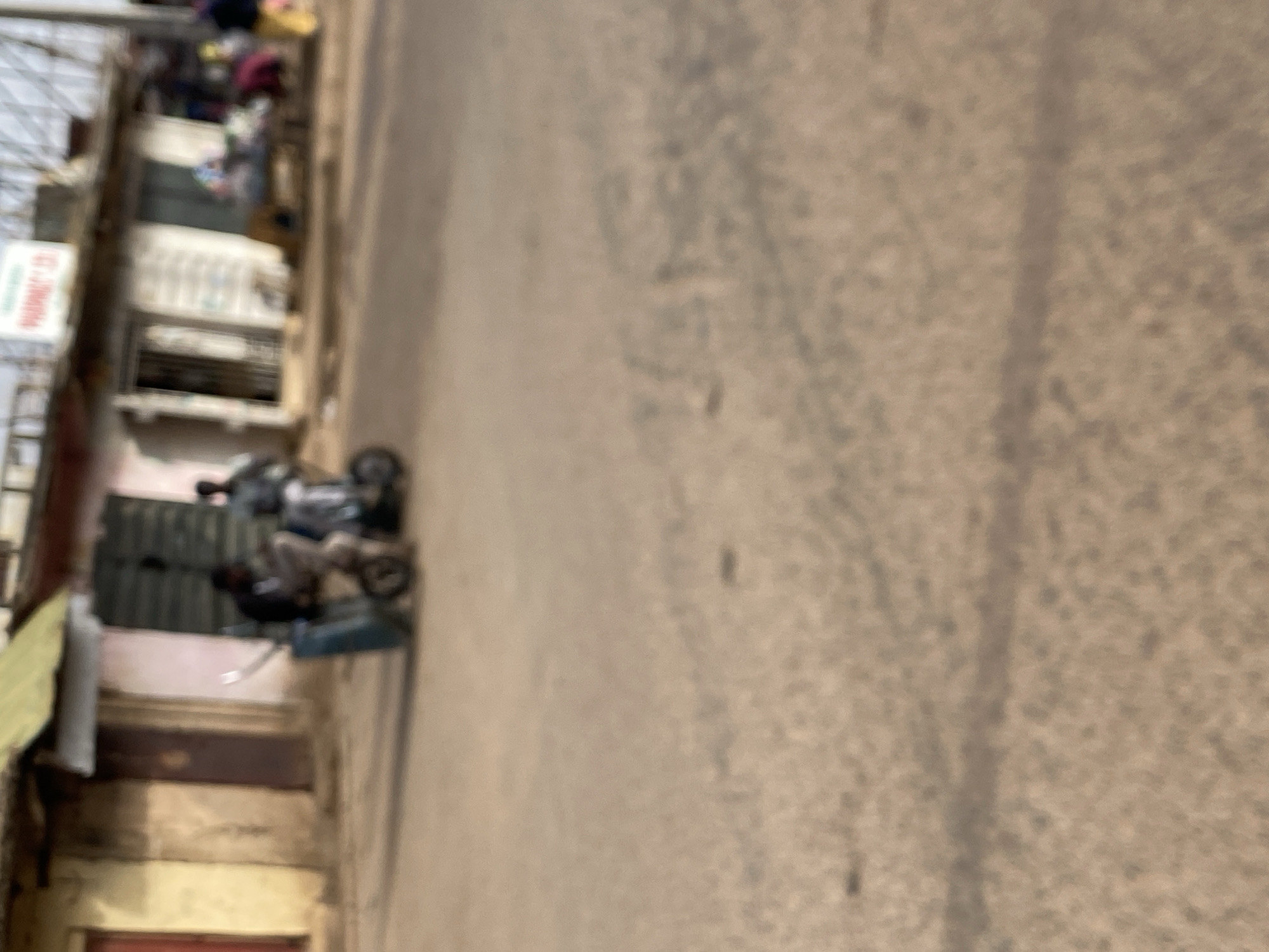 The scene is a street in Bukuru, Nigeria. A building lines the left side of the image, its storefronts displaying goods and signage. A motorcycle is parked near the building. The street itself is a light, speckled, textured surface. The photo is blurry.