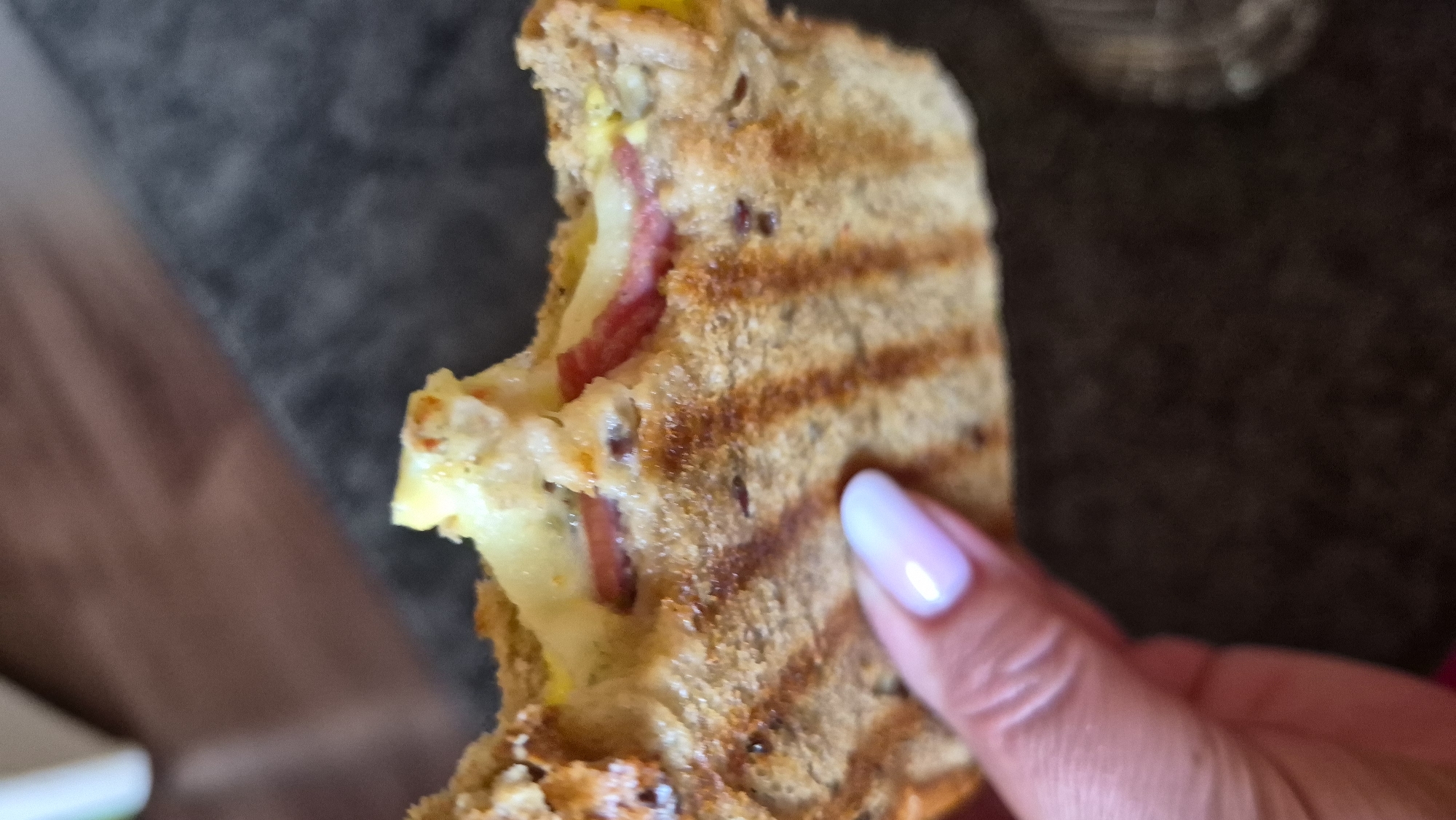 A close-up shot shows a person's hand, with a pale pink-colored fingernail, holding a half-eaten grilled sandwich. The sandwich has visible grill marks on its toasted bread and appears to be filled with melted cheese and some reddish-pink meat, possibly salami. The background is blurred, suggesting an indoor setting with a dark, textured surface. There is no discernible information about the location in Krauja, Latvia, in this image.