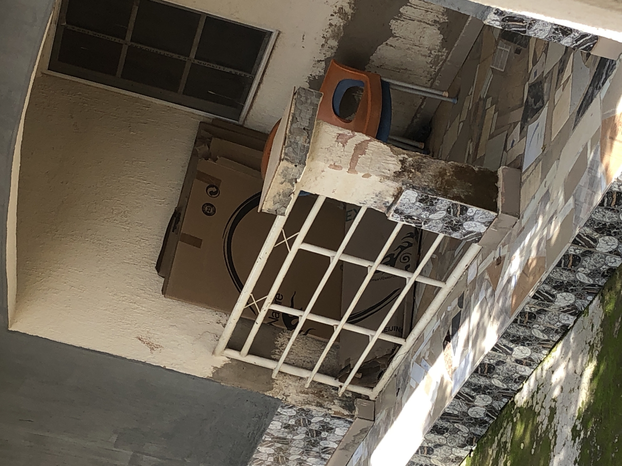 The image shows an outdoor scene, possibly a balcony or patio area, with various objects and architectural elements. A large cardboard box is prominently displayed, resting against a textured, cream-colored wall. Above the box, a window with a dark frame and divided panes is visible. To the right of the box, there is a partially constructed or damaged structure with a light-colored surface, adorned with black and white patterned tiles. A bright orange plastic chair with a blue seat is placed on this structure. A white metal grate or railing is positioned in front of the cardboard box. The background reveals a wall covered in a mosaic of rectangular tiles in various shades of brown, beige, and gray. The overall impression is one of an unfinished or neglected outdoor space. The lighting suggests it is daytime, with shadows indicating sunlight. There is some green moss or algae growing on the wall on the far right, hinting at a humid environment. Text is visible on the cardboard box, including circular logos and the word "Nature" and "BEIJING".
