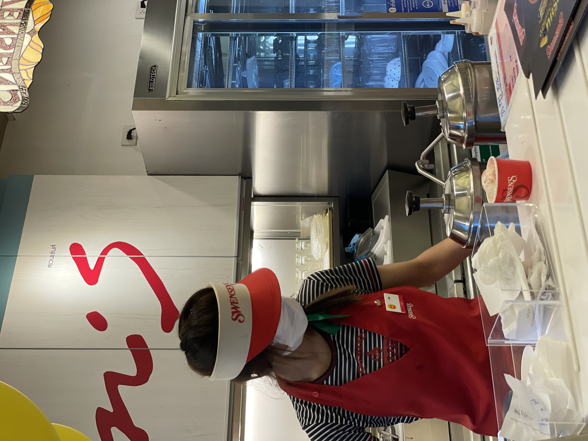 The image depicts a worker at a Swensen's ice cream shop in Pattaya City, Thailand. The worker, dressed in a Swensen's uniform (striped shirt and red apron), is scooping ice cream from a stainless steel dispenser into a red Swensen's cup. The worker is wearing a visor with the Swensen's logo and a face mask. The background includes a white wall with the Swensen's logo, an ice cream freezer unit labeled "KOLDTECH," and various containers and utensils related to ice cream service.