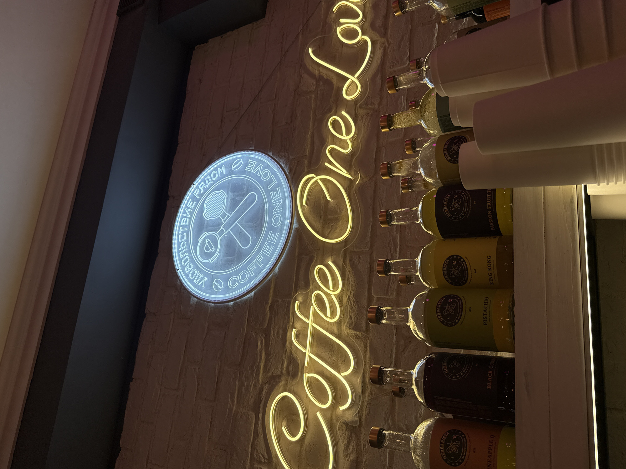 This media file shows a close-up, angled view of a coffee-themed display in what appears to be a cafe or bar. The primary subjects are neon signs and bottles.
On the left, a circular neon sign glows in white, featuring text that appears to be in Cyrillic script and English. The Cyrillic text reads "ДОВОЛЬСТВИЕ РЯДОМ" (Dovol'stviye ryadom), which translates to "Satisfaction Nearby". Inside the circle is a stylized graphic of a coffee portafilter and a coffee cup. To the right of this sign, a larger, bright yellow neon sign spells out "Coffee One Love" vertically.
To the right of the neon signs, a vertical arrangement of glass bottles is mounted on a white shelf. The bottles contain colorful liquids, and some have labels with text. Visible labels include "HERBARISTA", "PISTACHIO", "BLACK CURRANT", "PINEAPPLE Q", "KING KONG", and "PASSION FRUIT FI". The bottles are stacked in rows, with white paper cups or containers placed on top of some of them. The background is a textured brick wall, illuminated by the neon lights and what appears to be a warm, ambient light.
The overall scene suggests an indoor setting, possibly a modern cafe or a retail space related to coffee or beverages. The time of day is not discernible, but the lighting creates a cozy and inviting atmosphere. The location context provided is Moscow, Russia. There are no people visible in the image.