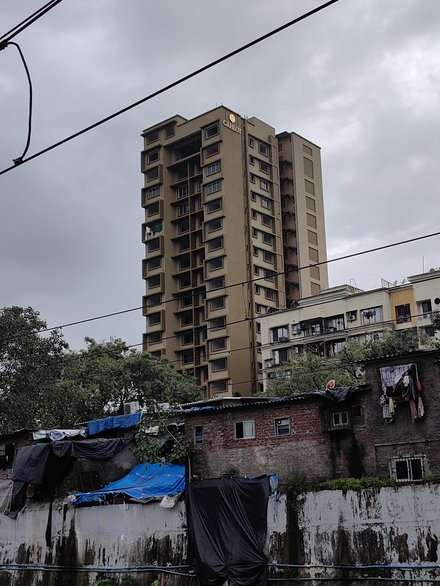 The image depicts a scene in Mumbai, India, characterized by a stark contrast between urban development and informal settlements. In the background, a tall, modern residential building with beige-colored walls and numerous rectangular windows dominates the skyline. The building features a prominent sign that reads "GAHLOT" with a hexagonal logo. In front of this building, a series of lower-rise structures, some appearing to be older apartments or commercial buildings, are visible, with air conditioning units and satellite dishes attached to their exteriors.
Closer to the foreground, a line of informal dwellings or shanties is present. These structures are characterized by makeshift roofs covered with tarpaulins in various colors, including blue and black, and walls constructed from brick and concrete. Some laundry is hanging outside these dwellings. A concrete wall with peeling paint and signs of weathering runs along the bottom of the frame, separating the informal settlement from what appears to be a lower area, possibly a drainage channel, with some greenery. Power lines are strung across the upper portion of the image, indicating the presence of infrastructure. The sky is overcast, suggesting a cloudy or possibly rainy day. The overall impression is one of a densely populated urban environment where different socioeconomic strata coexist, marked by the juxtaposition of modern high-rises and informal housing.