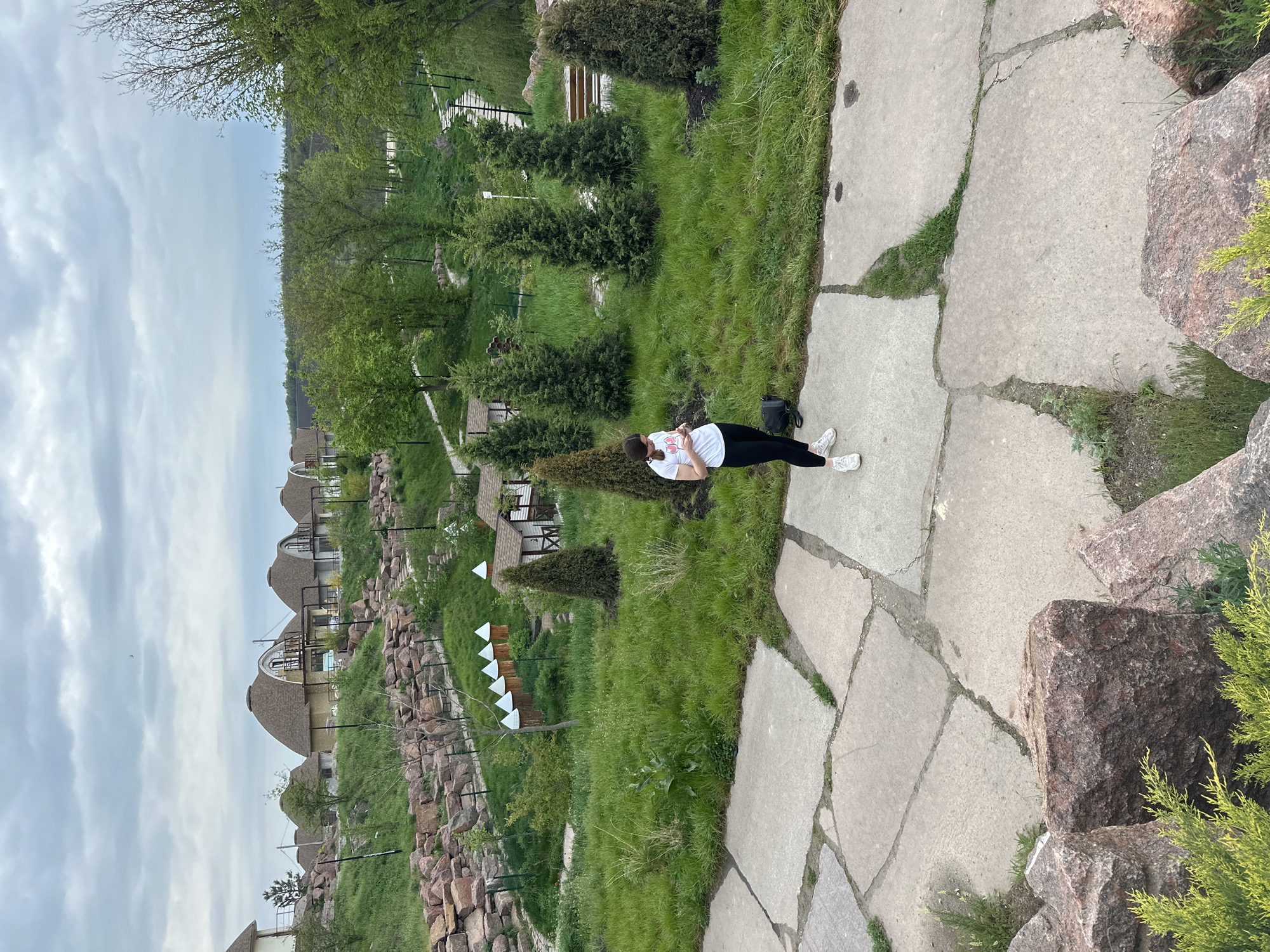 A woman stands on a concrete path in a landscaped area, possibly Zakharivka, Ukraine. She wears a white t-shirt, black pants, and white sneakers. A black bag sits near her feet. The path is constructed of irregularly shaped concrete slabs. To the left, a stone wall borders a grassy area with manicured evergreen trees and small, peaked-roof structures. In the background are buildings with rounded roofs. The sky is overcast.