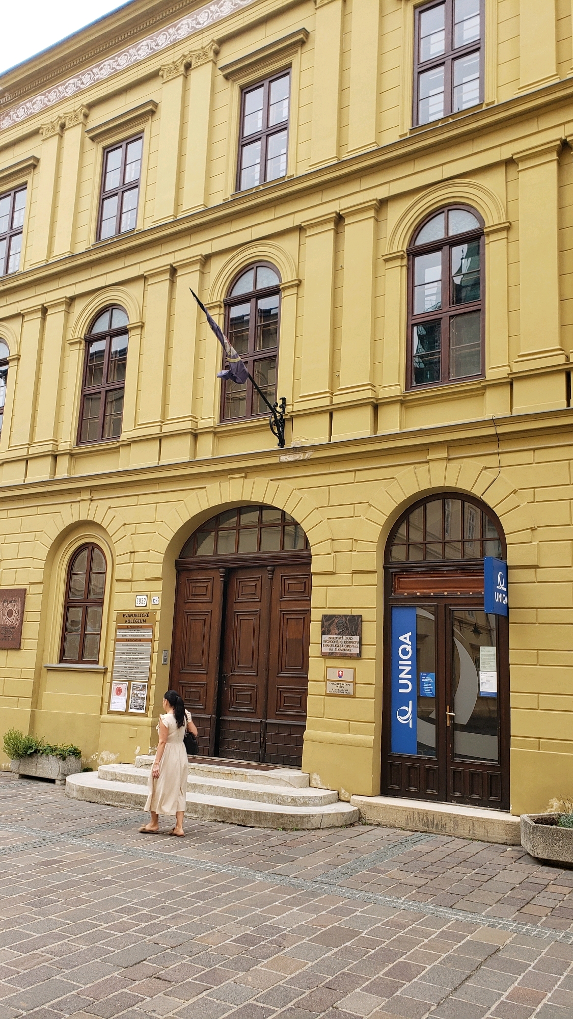A woman in a light-colored dress stands on a stone-paved street, facing a large, ornate wooden double door of a yellow building. The building has arched windows with dark frames, and a flag is mounted on the wall above the door. There are plaques on the building's facade, including one with text in Slovak and another with the "UNIQA" logo. The street is paved with cobblestones, and there are planters with greenery visible. The scene appears to be in the city of Prešov, Slovakia, based on the presence of Slovak text on the building's signage.
