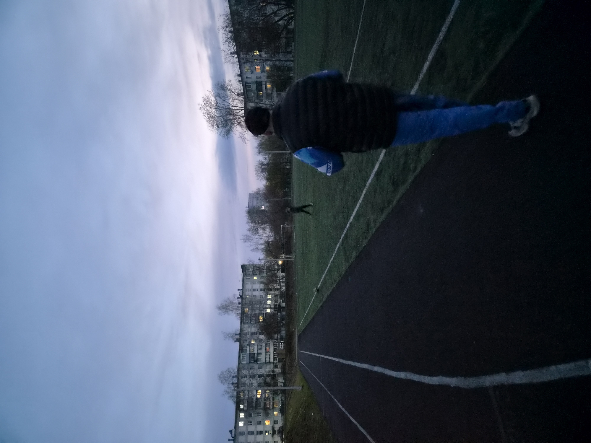 A tilted, eye-level shot shows a person on a running track, likely in Chaikovsky, Russia, based on the architectural style of the apartment buildings in the background. The person is wearing dark outerwear, blue pants, and light-colored shoes. They are standing near a grass field marked with white lines. A running track is visible, delineated with white lines. The sky is overcast with a blue-grey hue. In the background, apartment buildings are visible with lit windows. A single person stands in the center of the grass field at a distance from the camera. The image is shot at dusk or dawn, and the subject is the person standing on the running track.