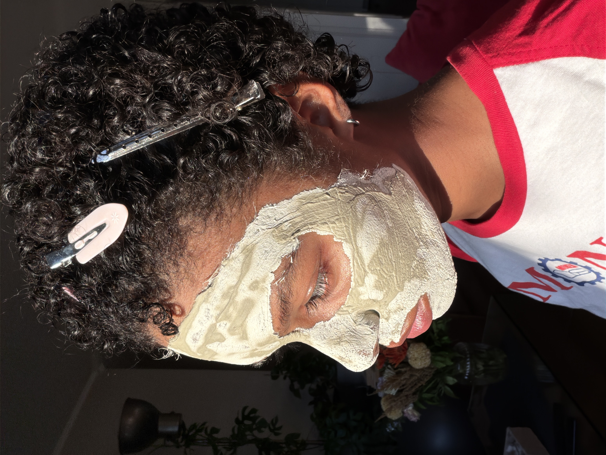 The image depicts a side profile of a person with curly dark hair, which is partially held back by two hair clips, one pink and one clear. A light-colored clay mask covers the person's face, obscuring much of their skin. Their eyes are closed, and their lips are parted slightly, revealing a hint of red lipstick. The person is wearing a white t-shirt with red sleeves and a red collar, featuring a blue and red logo with the text "MON." The background is dimly lit and shows a dark wall, a plant in a pot, and a glimpse of a bouquet of dried flowers in a glass vase on a dark surface. The scene is illuminated by strong natural light casting shadows. There is no specific indication of the location within the city of Champs-sur-Marne, France.