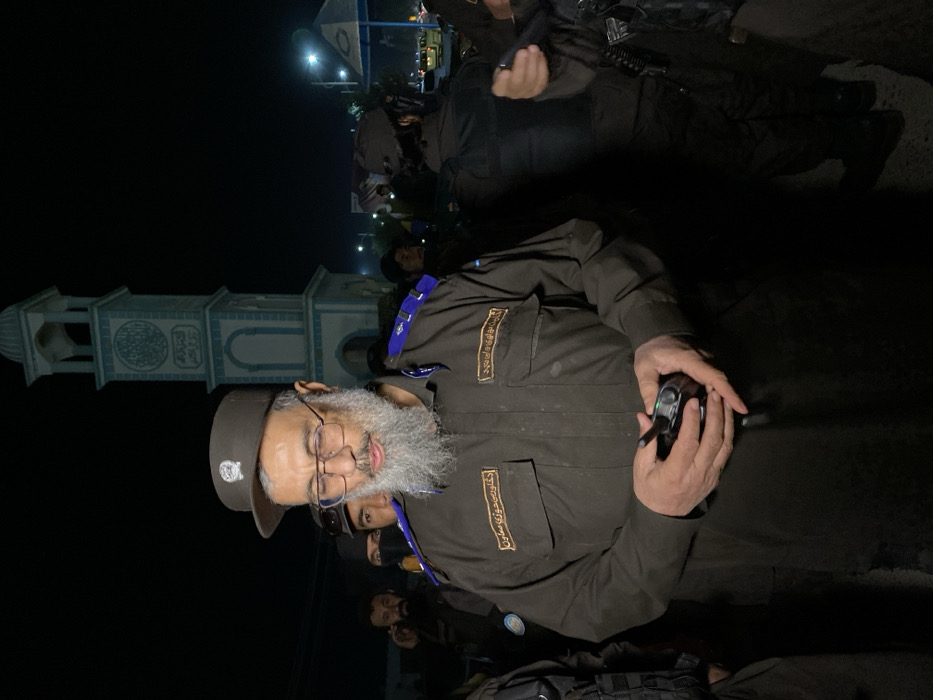The image captures an urban night scene in Kandahar, Afghanistan, featuring the Aino Mina Clock Tower. A man with a full grey beard and dark-rimmed glasses, wearing a dark olive-green uniform with blue shoulder boards and a dark baseball cap, occupies the foreground. Two patches with white Arabic script are visible on his chest pockets. He holds a black two-way radio with a green indicator light illuminated, his gaze directed downwards.
In the immediate background, several other men in dark uniforms are present, some partially obscured. One individual wears a black face covering. Further in the background, the distinctive Aino Mina Clock Tower, a multi-tiered structure adorned with light blue and white tiles and Arabic calligraphy, is illuminated against the dark sky. Artificial street lighting is visible in the distance, along with indistinct figures and faint vehicular traffic, indicating a city street at night.