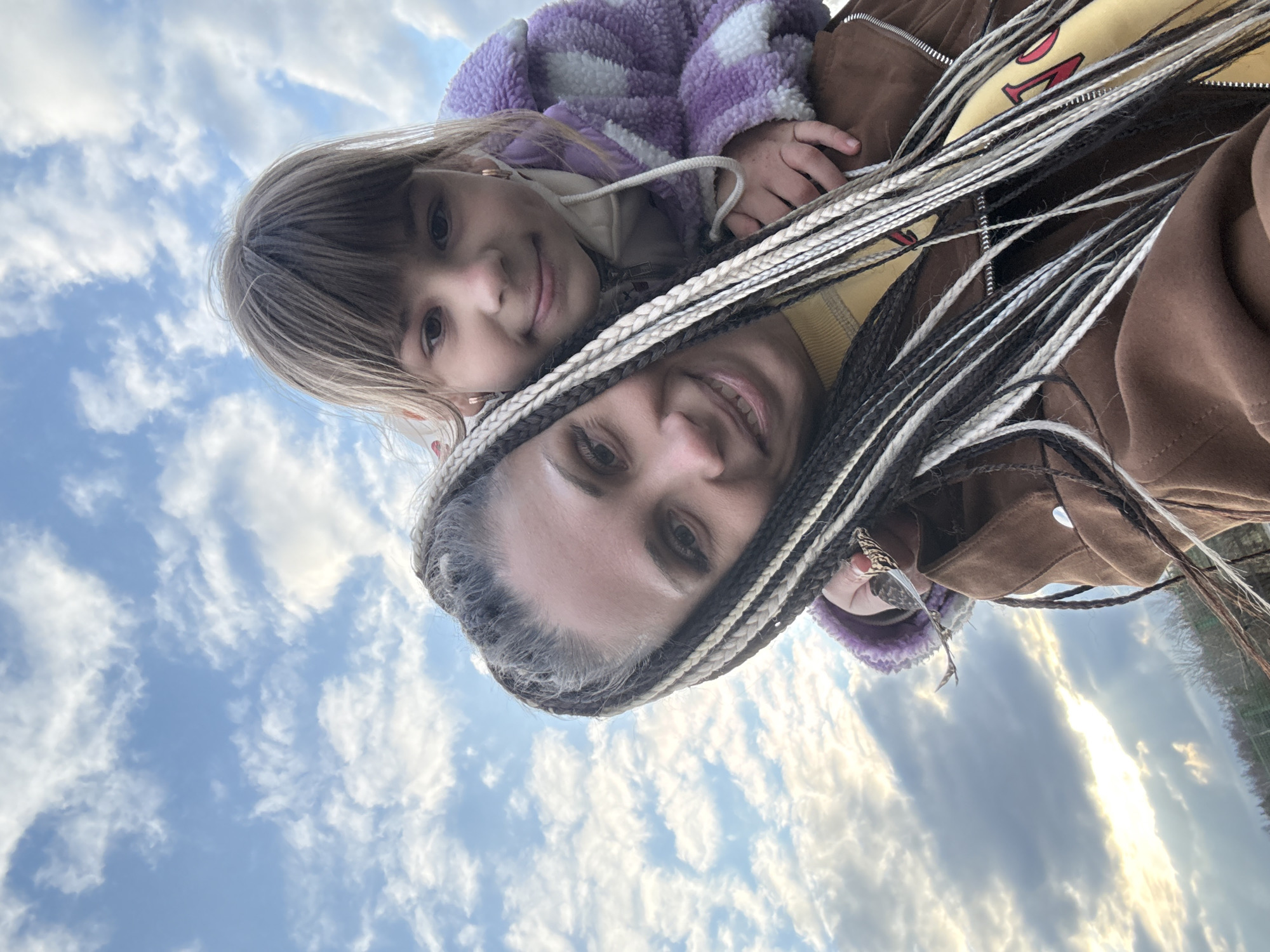 The image shows two people, a woman and a child, taking a selfie against a sky background. The woman, who appears older, is at the bottom of the frame, smiling. She has light skin and braided hair with alternating dark and light strands. The child is positioned above the woman, also smiling. They have fair skin and straight, light brown hair with bangs. The child is wearing a purple and white checkered jacket. The woman is wearing a brown jacket and yellow sweater. The background is a cloudy sky with a blue hue. The location is an outdoor area.