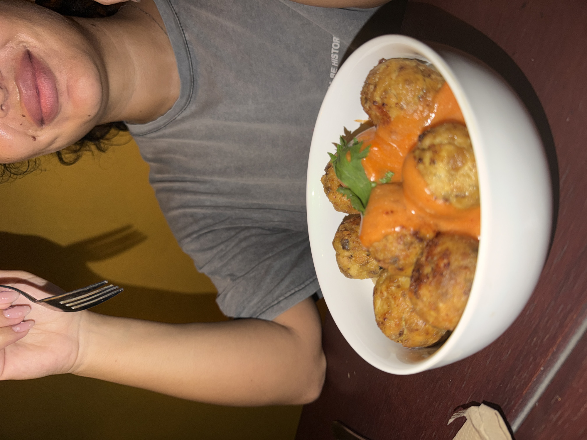 A close-up shot captures a bowl of meatballs in an orange sauce, garnished with a sprig of parsley, set against a dark wooden table. The person's hand, adorned with a light pink nail polish, holds a fork, poised above the food. The background features a muted yellow wall. The image appears to have been taken in North Kuta, Indonesia.