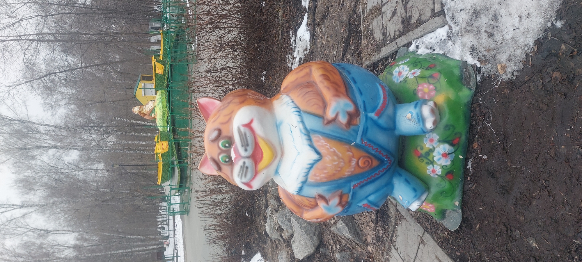 The image shows a whimsical garden sculpture of a cartoon cat dressed in a blue outfit and seated on a green, flower-painted base. The statue is located outdoors, near a patch of dirt and snow. In the background, bare trees and a fenced-in playground area with colorful structures are visible, suggesting a park or public space. The photo was taken in Novosibirsk, Russia.