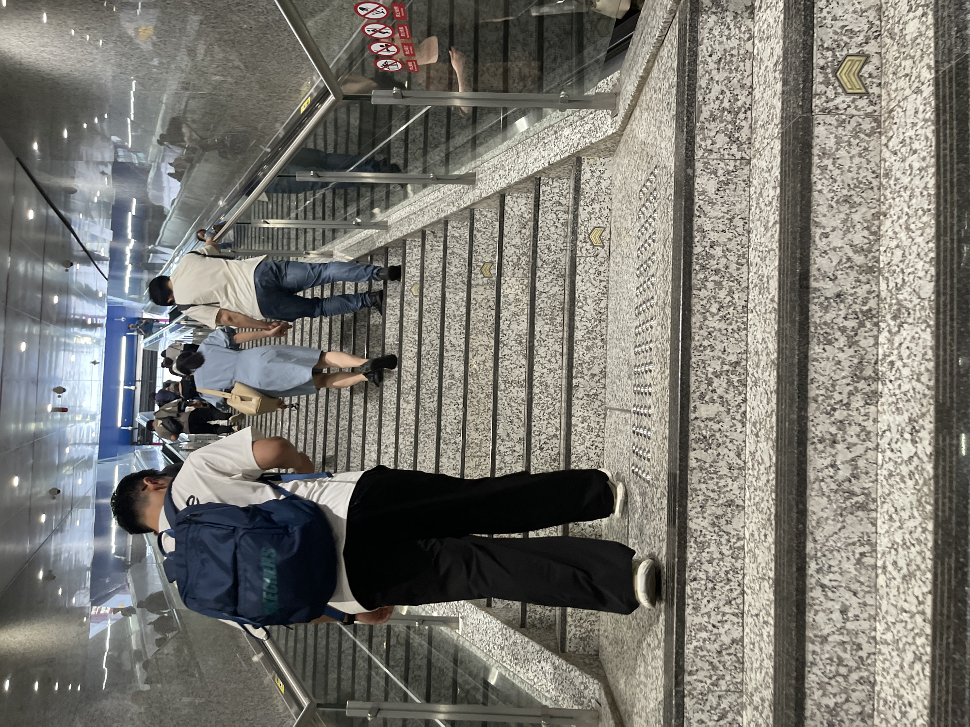 A group of people are ascending a wide, granite staircase in what appears to be a subway station in Panyu District, China. The man in the foreground, wearing a white t-shirt and black pants, carries a dark blue backpack. A woman in a light blue dress walks ahead of him, carrying a tan handbag. Another man, in a white t-shirt and blue jeans, follows them. The staircase has metal handrails and glass barriers. The surrounding environment is modern, with tiled walls and overhead lighting, typical of public transportation hubs. Several "no entry" signs are visible on the glass barrier.