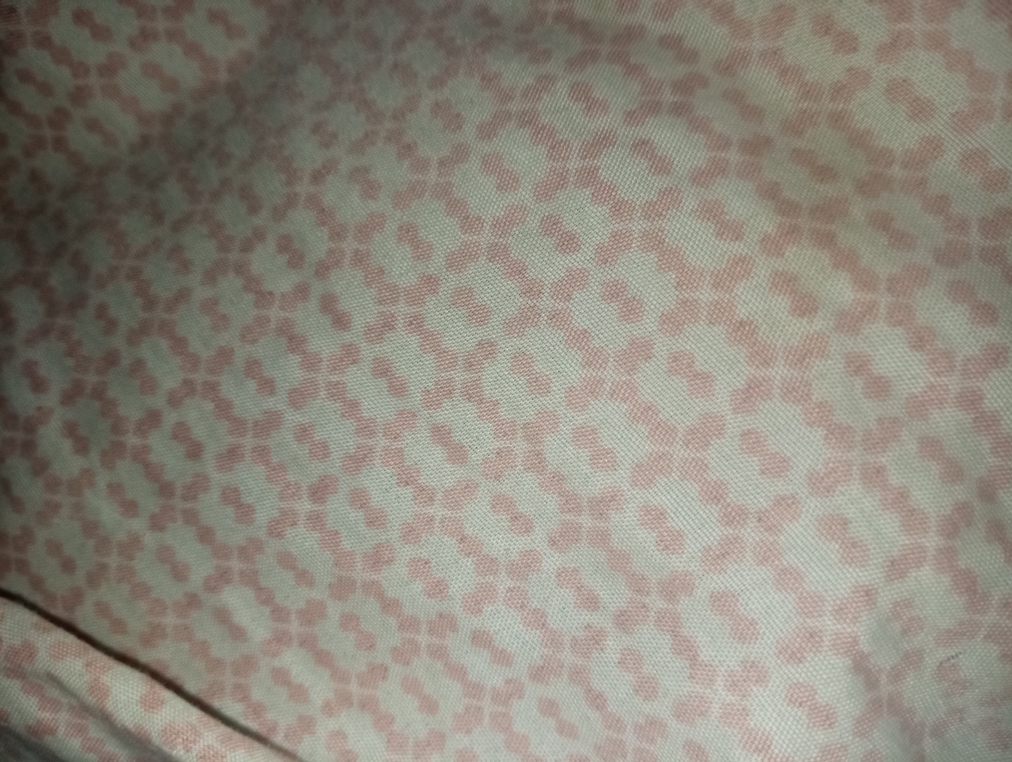 The image displays a woven textile surface in a city of Unknown, Unknown. The fabric has an off-white or light beige base color and features a repeating geometric pattern of small, solid pink shapes. Each pink shape consists of two connected ovals, resembling a barbell or dumbbell, oriented diagonally across the surface. These patterned elements are arranged in an offset grid, creating a dense, interlocking design. A visible diagonal fold or crease runs through the upper left quadrant of the fabric. The texture suggests a fine weave, possibly cotton or a synthetic blend. The lighting is soft and ambient, revealing the subtle texture and pattern without harsh shadows, although slight variations in brightness are present across the surface. No people, objects other than the fabric, or specific actions are depicted.