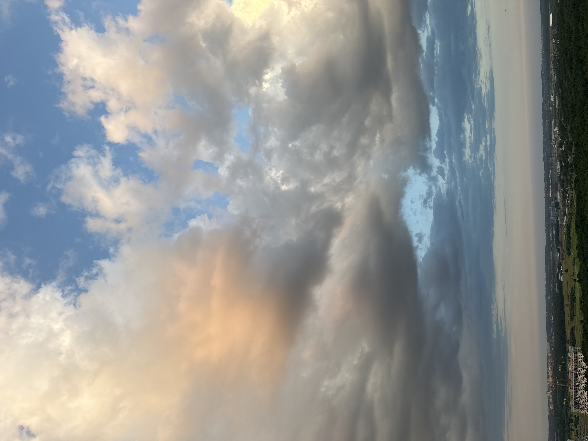 The image shows a sky filled with dramatic clouds over a landscape with buildings and trees. The clouds are a mixture of white, gray, and orange, suggesting a sunset or sunrise. In the distance, there is a city skyline with several buildings, some of which appear to be under construction. The foreground is dominated by a large, unfinished building with cranes. The overall scene suggests a view from a high vantage point, possibly a tall building or hill, looking out over the city of Lednevo, Russia. There are no people or animals visible in the image.