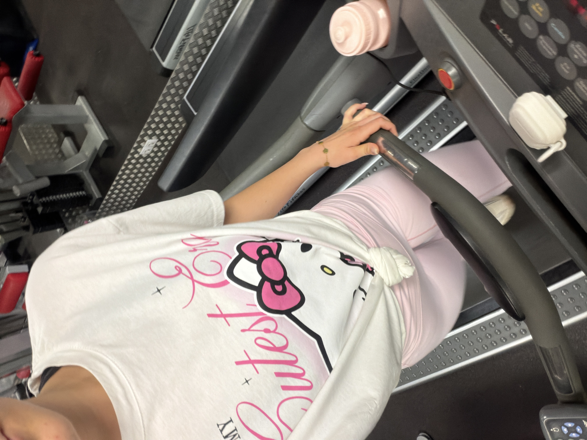 A person is standing on a treadmill. The person is wearing pink leggings and a white t-shirt with a Hello Kitty design. The person is holding onto the handrails of the treadmill. The treadmill is located in a gym. The location is unknown but possibly located in the city of Sofia, Bulgaria.