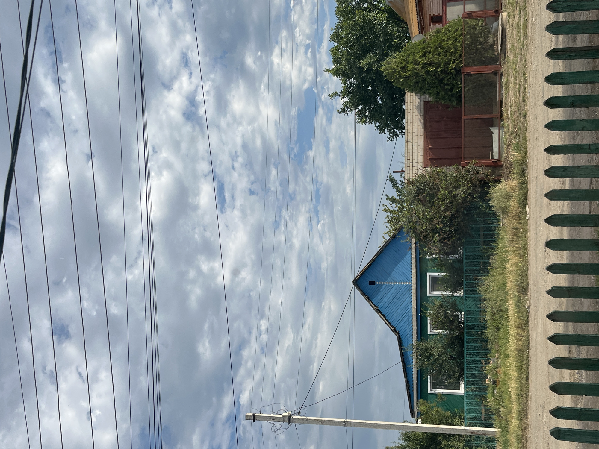 This image shows a rural scene in Krasnyye Barrikady, Russia. The foreground features a weathered green wooden fence. Beyond the fence, a dirt path runs alongside a green wooden house with a blue metal roof. The house has several white-framed windows. The scene is bordered by overgrown grass and bushes. In the background, power lines crisscross a cloudy sky. Some trees are visible, including a large green coniferous tree near a brick building. The overall atmosphere is one of quiet, everyday life in a village setting. The lighting suggests it is daytime, likely overcast given the clouds. There is no visible text in the image.