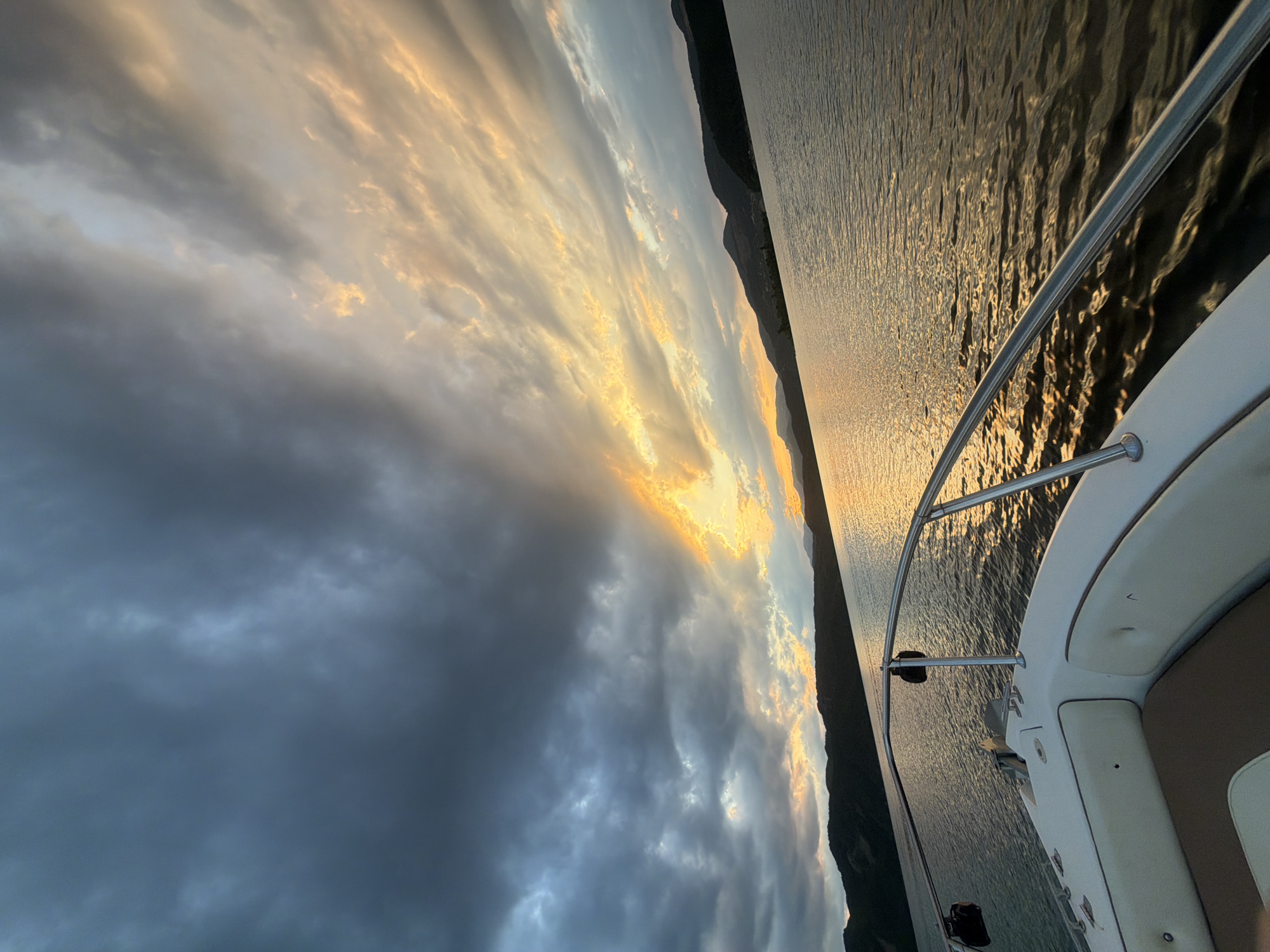 The image captures a scenic view from the bow of a white motorboat on a body of water, likely a reservoir, with a mountainous landscape in the background. The time appears to be either sunrise or sunset, given the dramatic lighting in the sky. Dark, heavy clouds dominate the left side of the sky, while the right side is illuminated by the sun breaking through, casting a golden and orange glow. The water's surface reflects the colors of the sky, creating a shimmering effect. The boat's railing and a portion of its seating are visible in the foreground. The location is identified as Glavatartsi, Bulgaria. There are no people visible in the image.