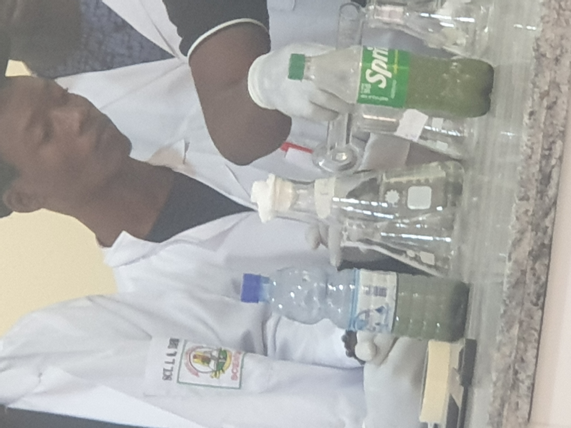The image shows a group of individuals in a laboratory setting, possibly performing a science experiment. One person is wearing a white lab coat with a logo that reads "SCLL B D". Another person is wearing a white lab coat and is holding a clear plastic bottle containing a green liquid and a blue cap. To the right, a green Sprite bottle with a green cap, also containing a green liquid, is visible. Several glass beakers and flasks are also present on the counter. The background appears to be a light-colored wall. The overall scene suggests a practical science or educational activity.