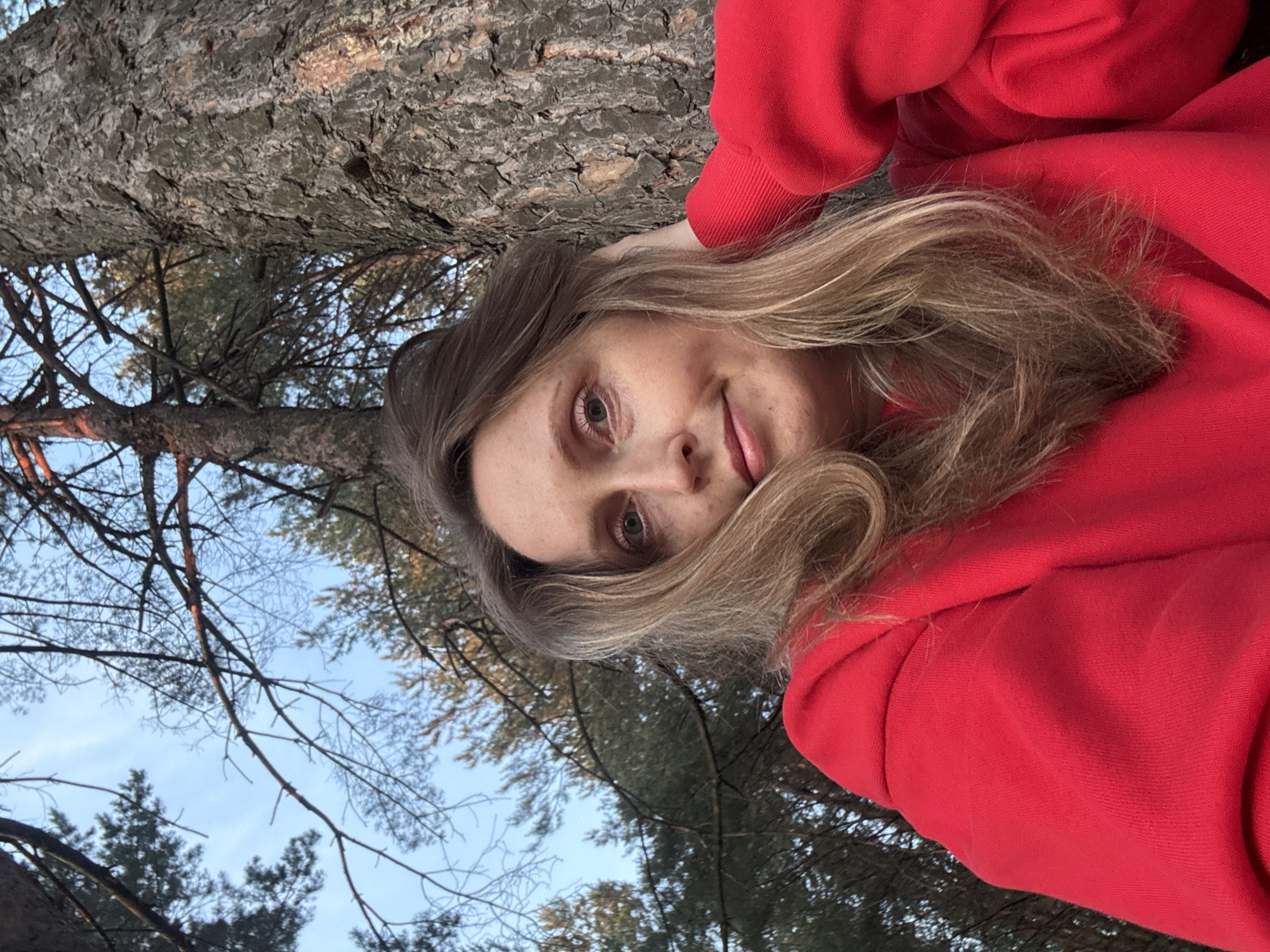 A medium-shot selfie is taken from a low angle of a woman with blonde hair wearing a red sweatshirt, leaning against a large tree. The woman is positioned on the right side of the frame, with her right hand supporting her head, and her left hand resting on her sweatshirt. She is looking towards the camera with a slight smile and her eyes are open. The background consists of trees and the sky, indicating an outdoor setting. The scene appears to be located in the city of Chaikovsky, Russia, based on the context provided. The lighting suggests it might be late afternoon or early evening.