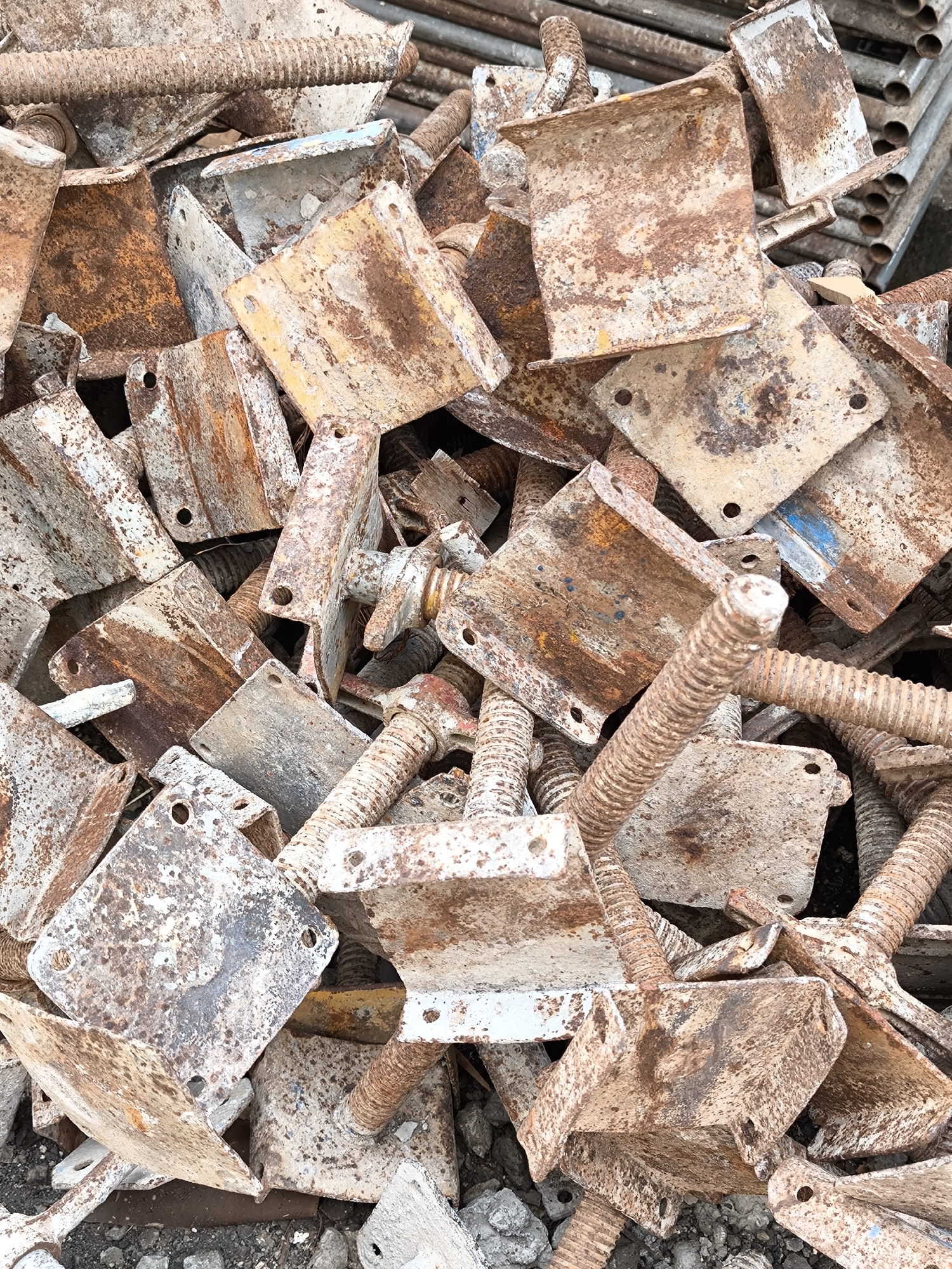 The image shows a close-up view of a pile of rusty and weathered metal construction materials. The objects consist primarily of adjustable screw jacks with square or rectangular base plates. These jacks are heavily corroded, showing rust and discoloration. The environment is cluttered and suggestive of a construction or demolition site in Unknown, Unknown. The focus is on the aging and used condition of the metal components.