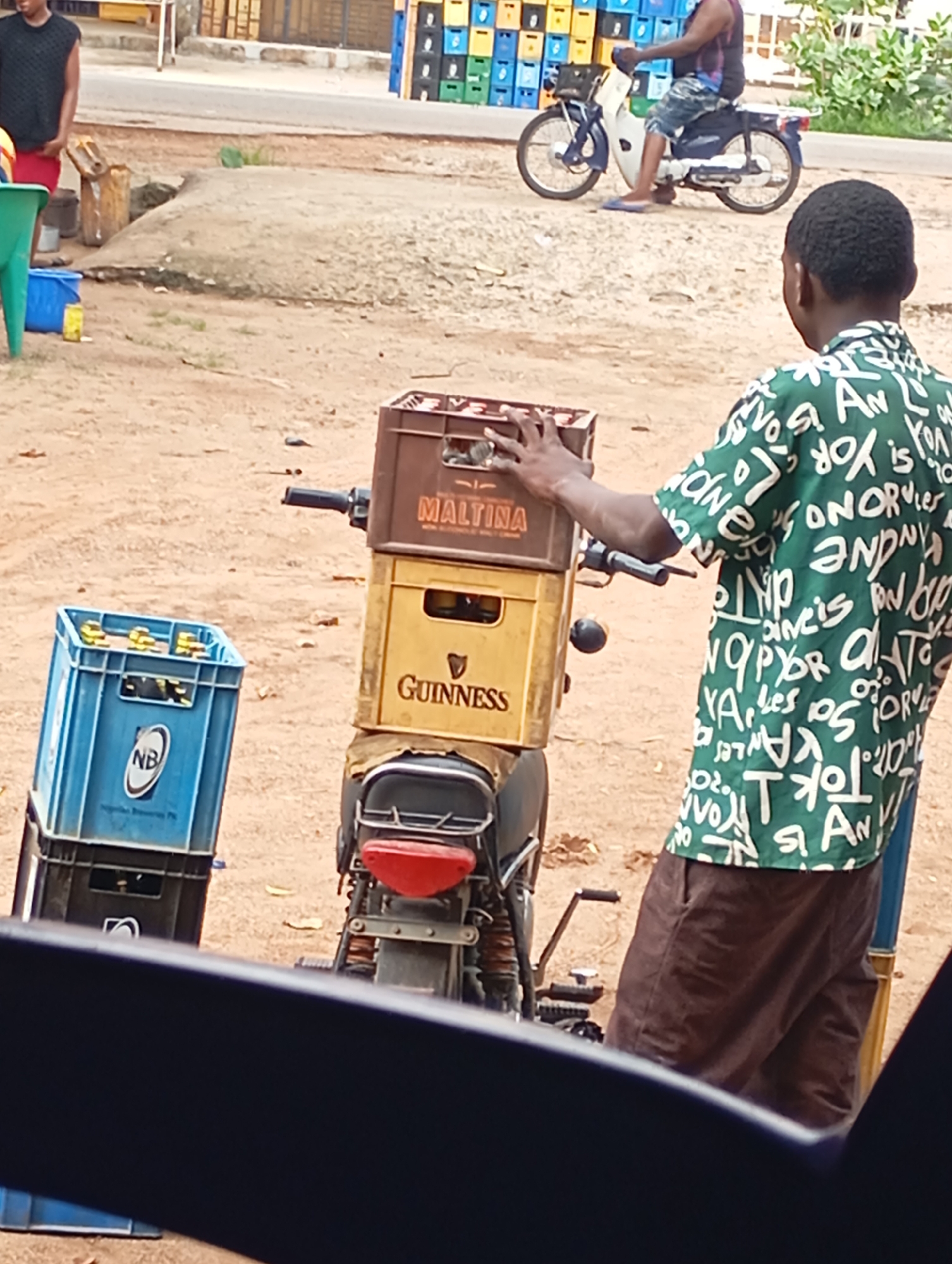 The image shows a street scene in Shendam, Nigeria. In the foreground, a young Black man wearing a green shirt with white lettering and brown shorts stands beside a motorcycle loaded with crates of beverages. The motorcycle has a red taillight and a black seat. On the motorcycle, there are two crates stacked on top of each other. The top crate is brown with "MALTINA" printed on it, and the bottom crate is yellow with "GUINNESS" printed on it. To the left of the motorcycle, there is a blue crate labeled "NB" containing bottles. In the background, another Black man is riding a white and blue motorcycle, and behind him are stacks of colorful plastic crates. The ground is sandy and dirt. The scene appears to be an informal market or distribution point for beverages.