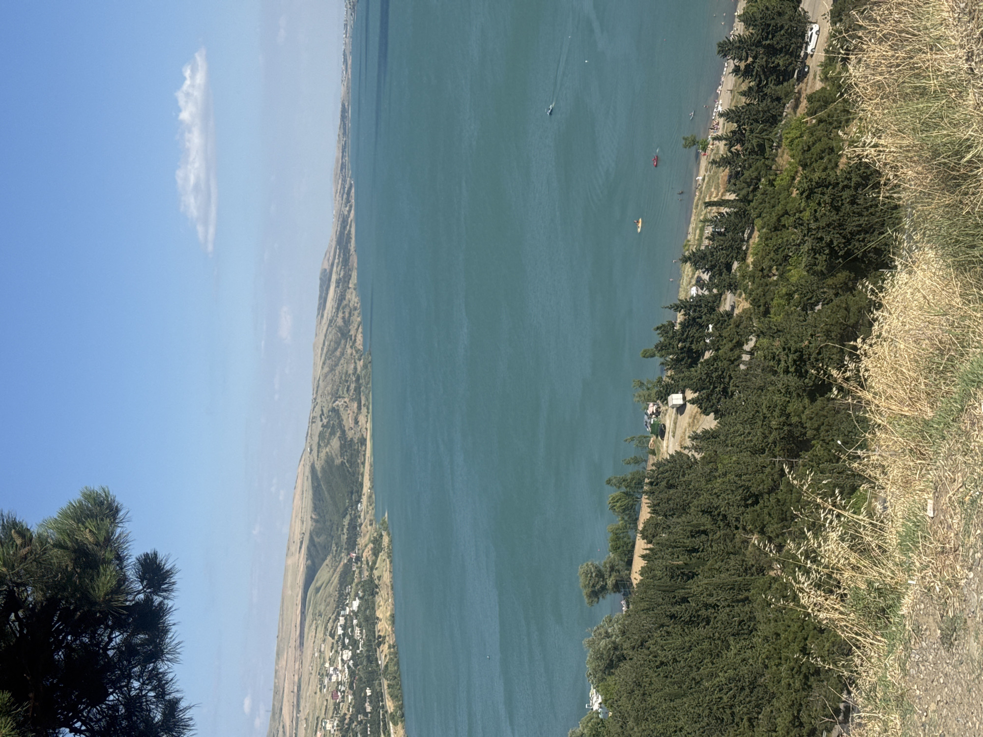 The image shows a scenic view of a large body of water, likely a reservoir, bordered by hills and a developed shoreline. On the water, several kayaks or small boats are visible, suggesting recreational activity. The shoreline features a road with parked cars and people gathered, indicating a populated area. Lush green trees line the edge of the water, and the hills in the background are covered in sparse vegetation. The sky is blue with a few wispy clouds. The scene appears to be set on a sunny day, likely during daytime. The perspective is from a high vantage point, looking down at the water and the surrounding landscape.
