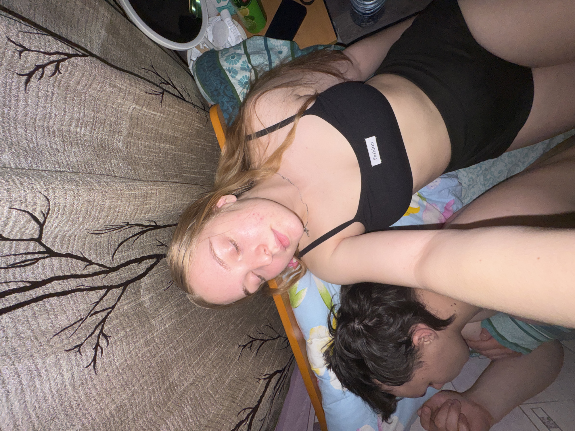 The image captures a close-up, high-angle interior scene in the city of Perm, Russia, depicting two individuals in a bed. In the foreground, positioned centrally and to the right, a young blonde female is visible from the waist up. She has fair skin with visible blemishes on her cheek and forehead, and her eyes are closed. Her blonde hair is parted on the side and extends down her back. She wears a black spaghetti-strap crop top with a white rectangular "Fashion" label centered on the chest, and black shorts. Her left arm is extended towards the bottom left, holding the camera, suggesting a self-portrait.
To her left, slightly lower in the frame, a young male is lying on his right side, facing away from the camera. He has dark, short, curly hair and fair skin. His right arm is bent, with his fist near his head, and he appears to be asleep, partially covered by a light blue and white striped blanket or sheet. Both individuals are lying on patterned pillows and sheets featuring a light blue background with white and teal floral designs.
Behind the individuals, a light brown/beige curtain with a textured fabric and a dark, stylized bare tree branch pattern is visible. An orange-brown headboard or bed frame runs horizontally behind the female's head. To the far right, a cluttered bedside area includes a round silver-framed mirror, various bottles, a green object, a black rectangular item, and a stack of clear glasses. The lighting is dim, suggesting an indoor night setting.
