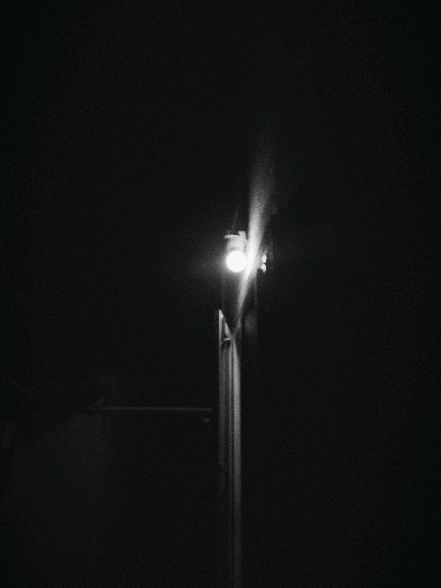 A black and white photograph captures a streetlamp at night in Nizampatnam, India. The lamp is illuminated, casting a soft glow into the darkness. The pole supporting the lamp is visible, as is a faint haze or smoke emanating from the light source, suggesting a misty or dusty environment. The background is predominantly dark, indicating the absence of other significant light sources and contributing to the overall nighttime atmosphere. A dark, indistinct structure, possibly a building or wall, is partially visible to the left of the lamp pole. The image has a grainy texture, typical of older film photography or specific digital processing.