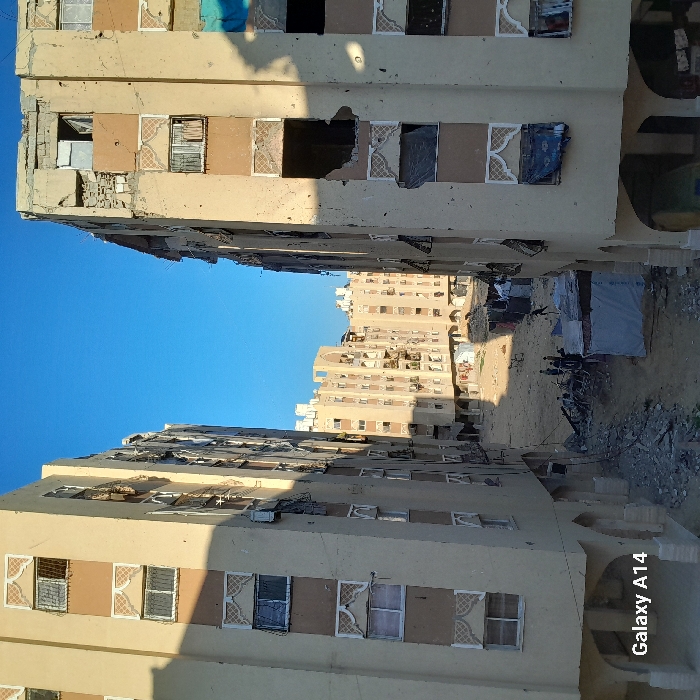 Buildings display significant damage, with broken windows and structural debris, potentially caused by conflict. The scene shows signs of displacement, with makeshift shelters and scattered belongings on the ground between the buildings. The sky is clear, contrasting with the destruction below. The general atmosphere suggests a war-torn urban area in need of reconstruction.
