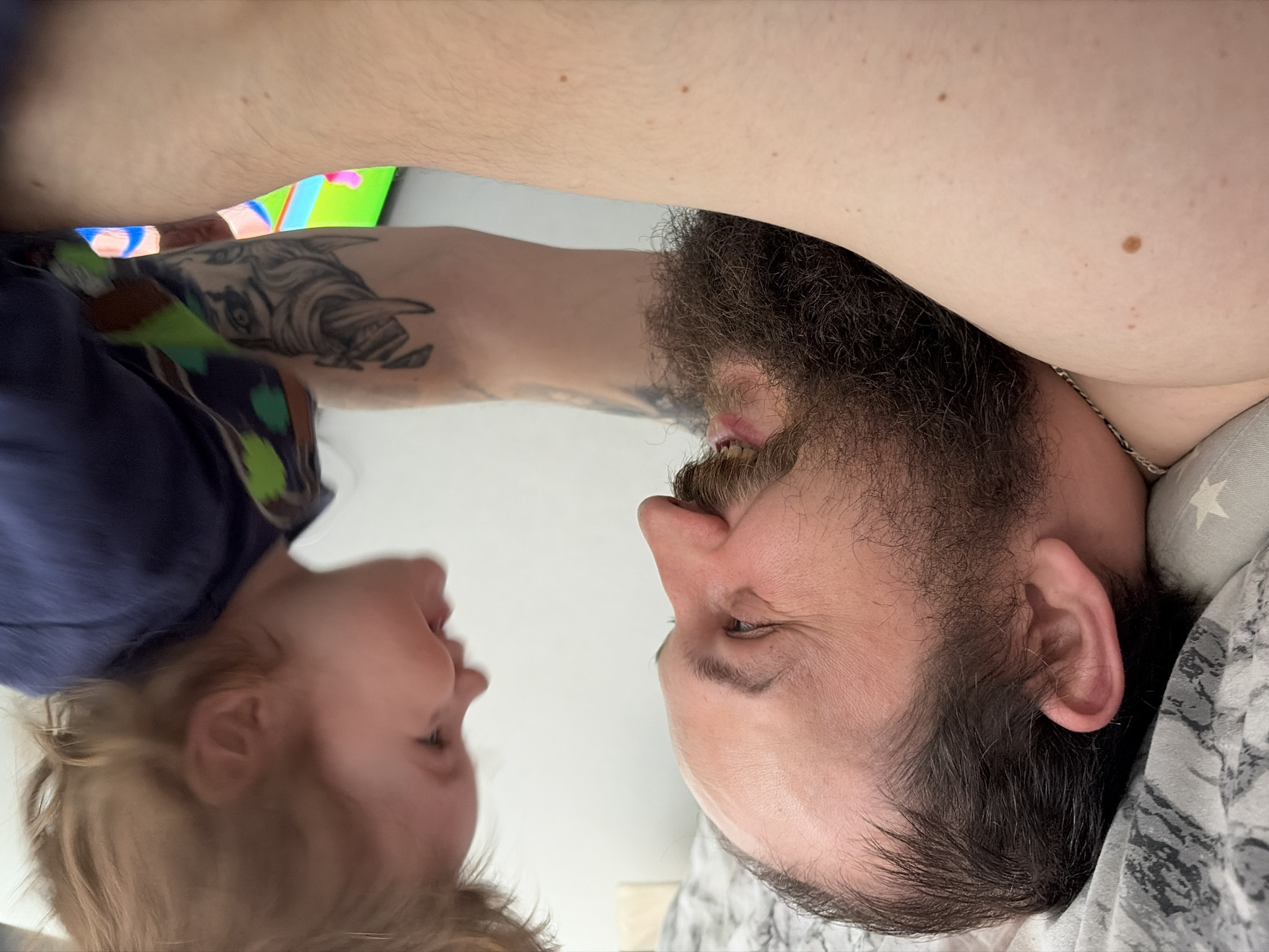 A man with a beard lies on a pillow with white stars. A fair-haired child faces the man. The man's arm with a tattoo extends over the child. In the background, a television shows a bright screen. The location is Chaikovsky, Russia.