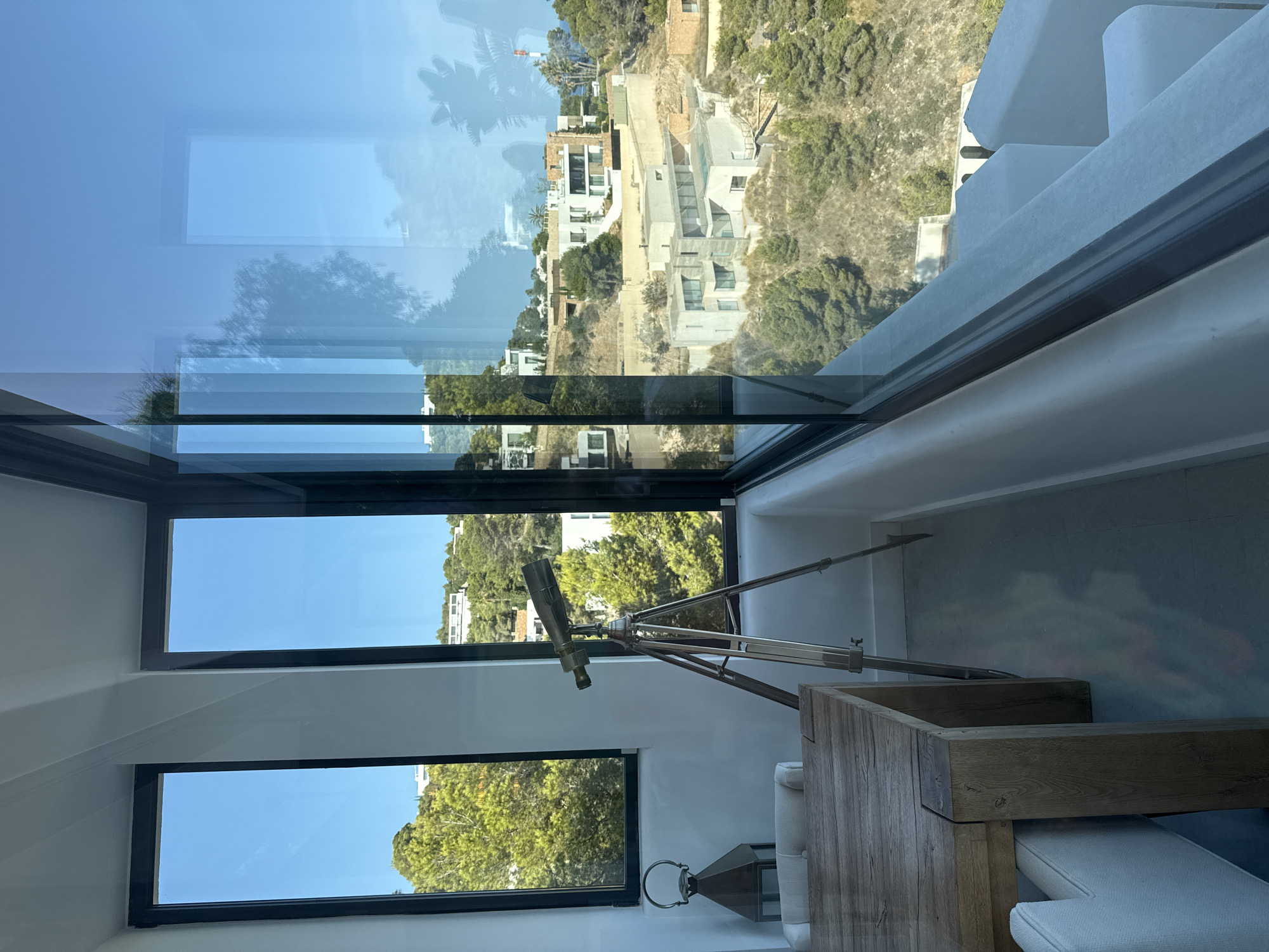 The image shows an interior view of a room with large windows looking out onto a hilly landscape with modern houses and trees. The room features a wooden dining table with white chairs and a vintage telescope on a tripod positioned near the window. A decorative lantern is also visible on the left side of the image. The view outside encompasses a sunny day with a clear blue sky and scattered clouds, showcasing the architecture of several multi-story residences nestled into the hillside, typical of the affluent coastal regions around Santa Eulària des Riu, Spain. The reflection in the glass shows a partial view of the interior space and the sky outside.