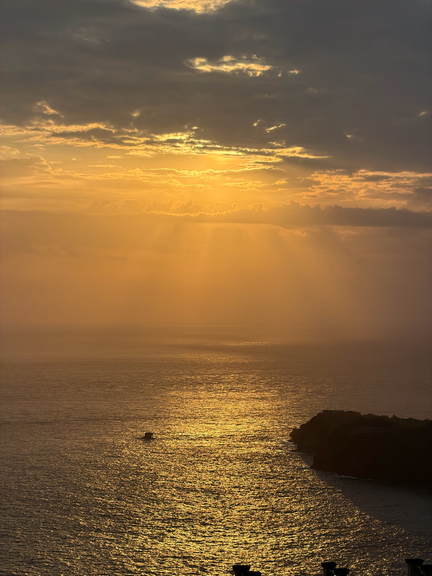 This vertical, wide shot captures a dramatic sunset over the ocean in Nusa Penida, Indonesia. The sky is a warm gradient of golden orange and diffused yellow, with scattered clouds. Sunbeams pierce through the clouds, creating a halo effect. The ocean below reflects the sunset, shimmering with golden light. In the foreground, a dark, silhouetted landmass juts into the sea on the right side of the frame. A small, dark object, possibly a boat or rock formation, is visible in the water to the left of the landmass. The bottom edge of the image shows the dark silhouettes of structures, hinting at a vantage point from a building or elevated area. The overall atmosphere is serene and awe-inspiring, dominated by the intense colors of the sunset.