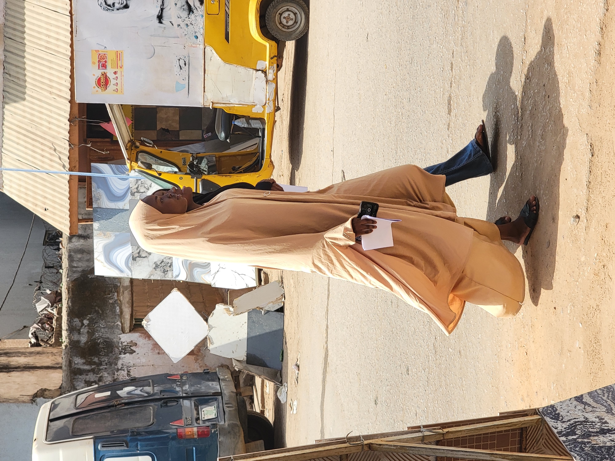 In Maiburiji, Nigeria, a person wearing a tan robe and blue jeans stands on a paved street, holding a black smartphone in one hand and a white piece of paper in the other. The person wears black sandals. The shadow of two people is cast on the street. Behind the person is a yellow auto rickshaw parked next to a white and blue van. There is a building partially visible with corrugated roofing and loose tiling.