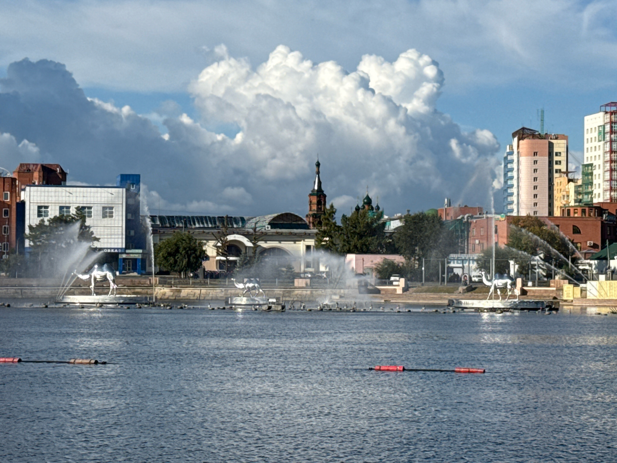 The image depicts a waterfront view of Chelyabinsk, Russia, specifically the area surrounding the monument "The Camel," located on the banks of the Miass River. In the foreground, the rippling surface of the river is visible, with two red buoys floating on its surface. In the middle ground, a circular fountain is active, with water jets creating a mist around a metallic sculpture of a camel. Several other smaller fountains are also visible along the embankment.
The background features a mix of architectural styles. To the left, a modern, multi-story white building with blue accents stands out, along with a red brick building adjacent to it. Further to the right, a row of modern high-rise apartment buildings, varying in color and design, dominates the skyline. Among these, a prominent white building with a red-striped facade is noticeable. Between the modern buildings and the river, older structures can be seen, including a building with a large arched entrance and a domed church spire. Trees are scattered along the embankment, and the sky is filled with dramatic, billowing white clouds against a bright blue backdrop, suggesting dynamic weather conditions. The overall scene captures an urban landscape with prominent water features and contrasting architectural elements under a vast, cloud-filled sky.