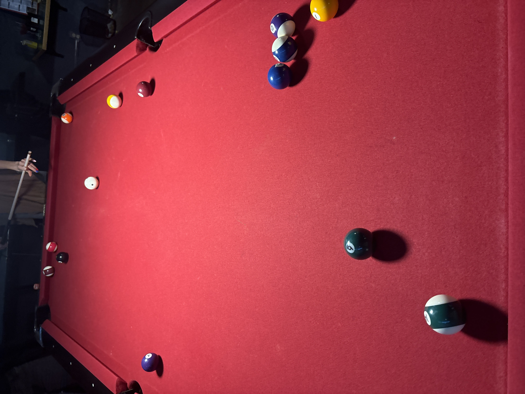 A high-angle indoor shot shows a red pool table with multiple billiard balls scattered across its surface. The balls are of varying colors and numbers, including solids and stripes. A hand holding a pool cue is visible on the left edge. The environment appears dimly lit with equipment. The specific location in Mexico City is unknown.