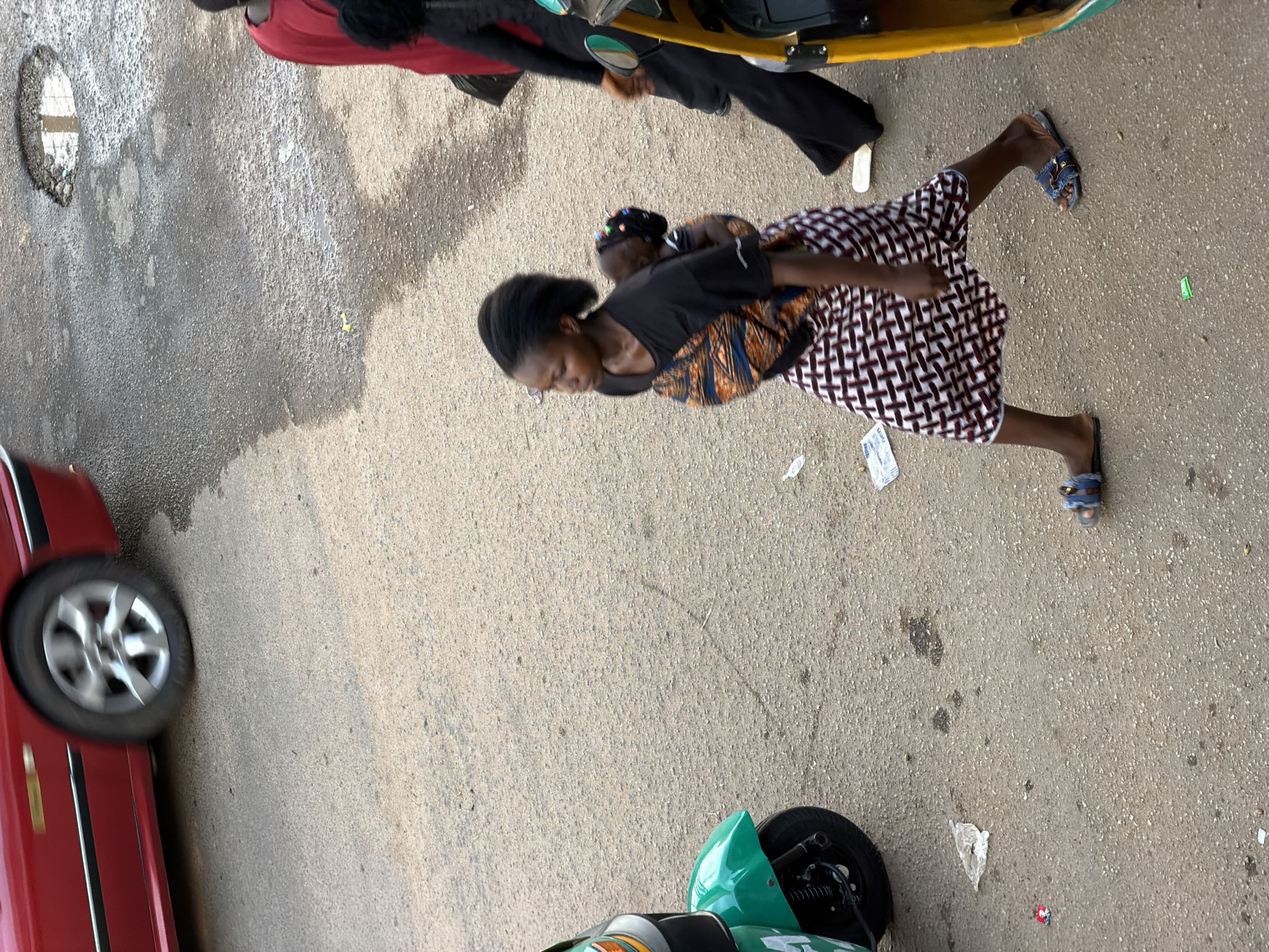 A woman is walking while carrying a baby on her back in Bukuru, Nigeria. The woman is wearing a black t-shirt with an orange and blue patterned wrap around her waist, and a black and white checkered skirt. The baby is wrapped in a colorful fabric, also on the woman's back. The ground is paved with asphalt and shows signs of wear and tear, with a few puddles and debris scattered around. In the background, a red car is parked, its wheel slightly blurred, indicating movement or a fast shutter speed. A green and white motorized tricycle is partially visible on the left side of the image, and a yellow and green vehicle is on the right. The lighting suggests it is daytime.