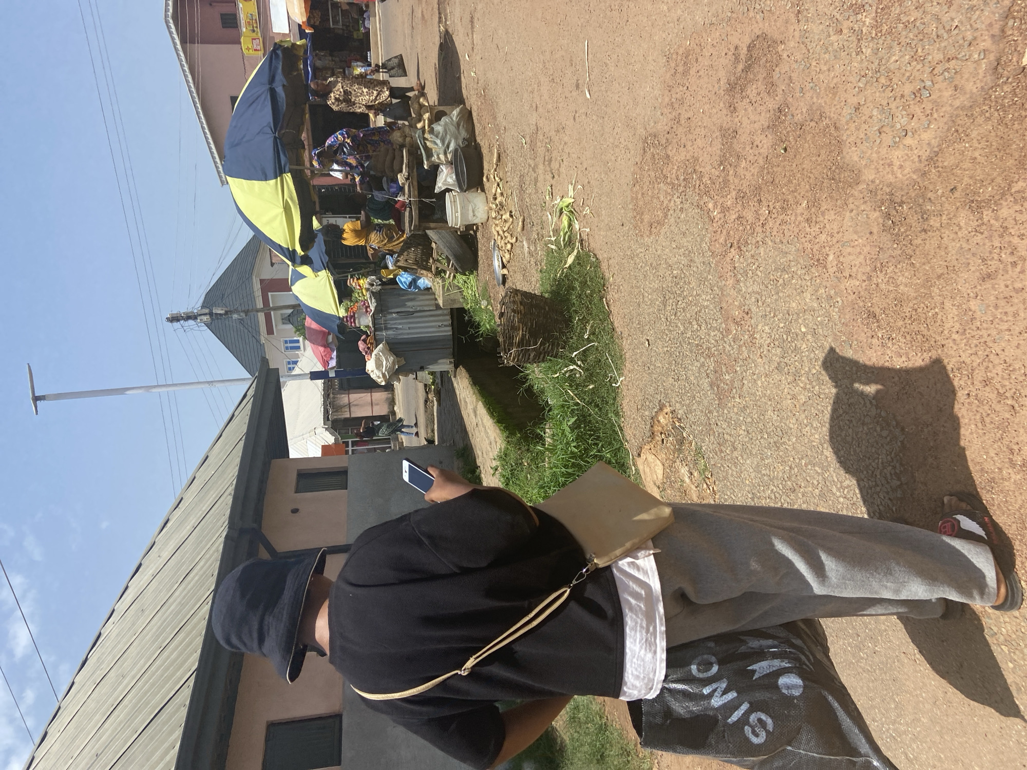 The image captures a street scene in Jos, Nigeria, on a bright, sunny day. In the foreground, a person wearing a black bucket hat and a black long-sleeved shirt is viewed from behind, holding a smartphone in their right hand. They are also carrying a large black shopping bag with white lettering on their left side and a cream-colored crossbody bag. Light gray trousers and dark sandals complete their attire. The ground is a mix of reddish-brown dirt and small pebbles.
In the mid-ground, a bustling market stall is visible under large blue and yellow umbrellas. Several people, primarily women, are engaged in selling and buying goods. Various items are displayed, including fruits, vegetables, tubers, and woven baskets. A metal bin and some sacks are also present. The background shows buildings with tin roofs and a street with other market stalls and pedestrians. Power lines crisscross the sky. The overall environment suggests a typical open-air market in a Nigerian city.