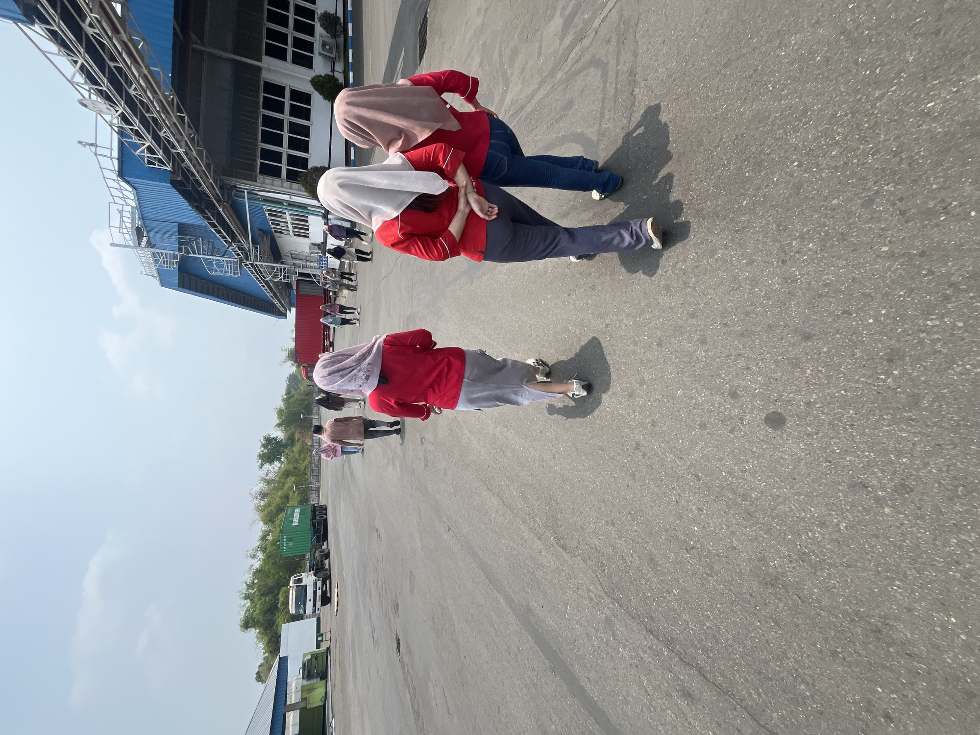 In Pelang, Indonesia, two individuals are walking away from the camera on an asphalt paved area. Both are wearing red long-sleeved shirts and headscarves. The individual on the left wears grey trousers and white sneakers. The individual on the right wears blue jeans and black and white sneakers. They are walking towards a blue and white industrial building with many windows. In the background, there is a white truck carrying a green shipping container. The sky is mostly clear with a few clouds. The scene appears to be a logistics or industrial facility.