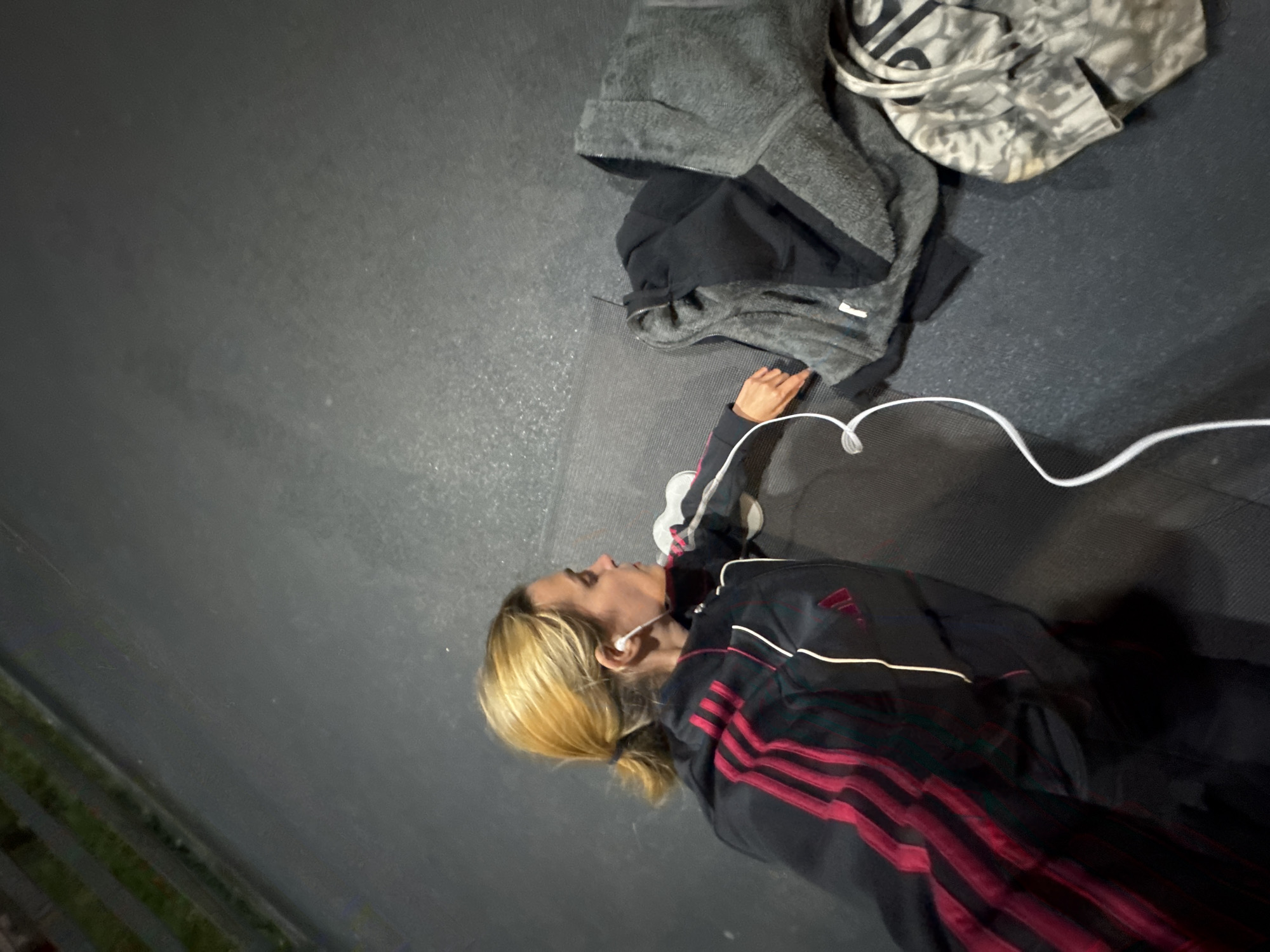 An overhead shot captures a person, likely a woman, lying on a dark grey surface, possibly an exercise mat or gym floor. She is positioned diagonally in the lower right portion of the frame, with her head towards the top right and body extending downwards. She has blonde hair pulled back in a low ponytail and is wearing white wired earphones in her ears.
She is dressed in an athletic jacket, dark in color, featuring prominent red stripes on the sleeves and body, consistent with the Adidas brand, whose logo is faintly visible on the jacket. Her eyes are open, and she appears to be looking upwards or slightly ahead, with a neutral expression, possibly resting or listening intently. Her right hand is visible, resting near her head.
To the upper left of her, several pieces of clothing are piled. These include a grey, fuzzy-textured hooded jacket, a black garment underneath it, and a lighter-colored, patterned item (possibly a bag or another piece of clothing) with a camouflage-like design. The lighting in the scene is dim, suggesting an indoor setting or nighttime. No specific location cues are visible within the image to indicate Tbilisi, Georgia, other than the provided context.
